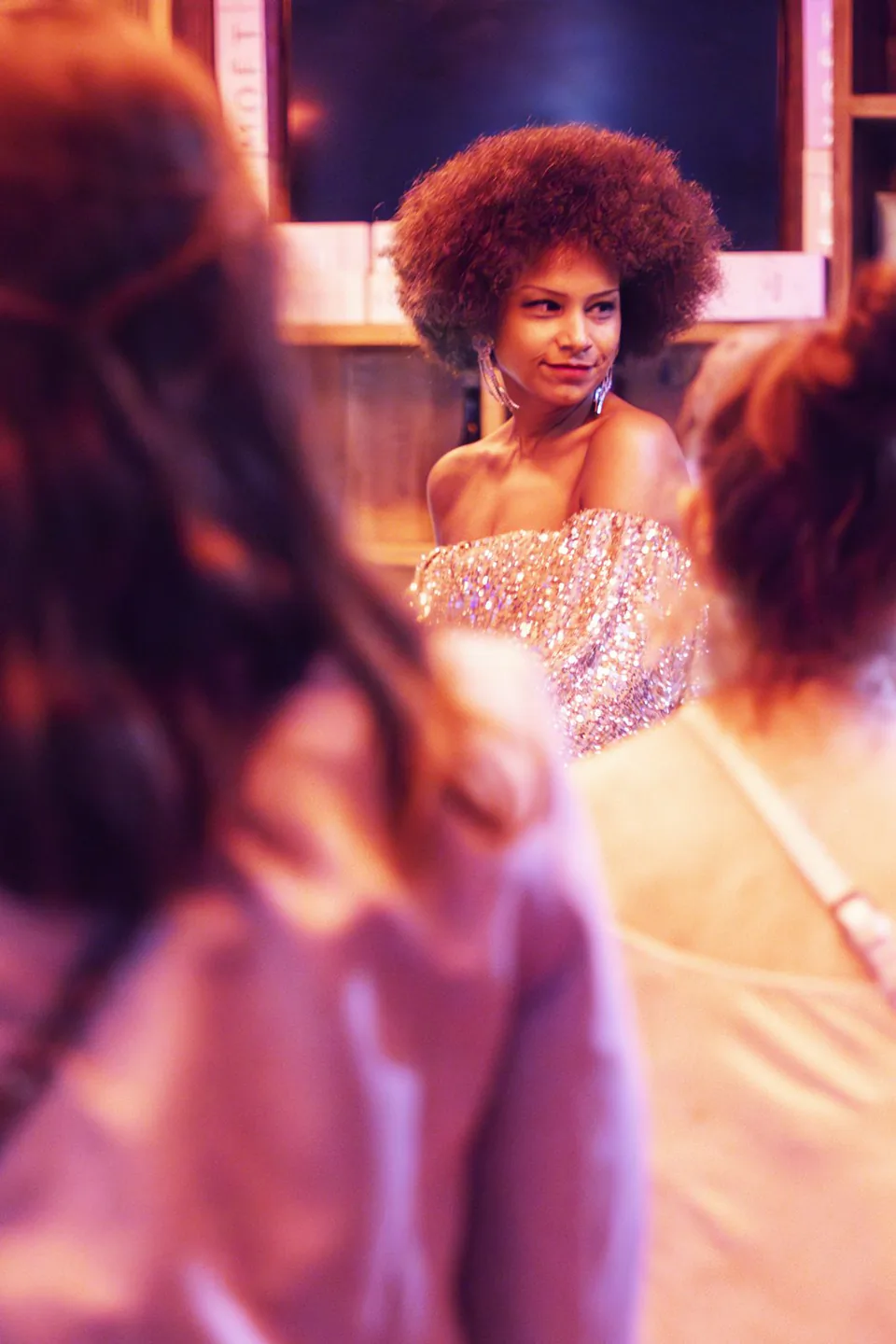 Frau mit Afrofrisur im glitzernden, schulterfreien Kleid zwischen Gästen bei warmem Eventlicht im Innenraum.