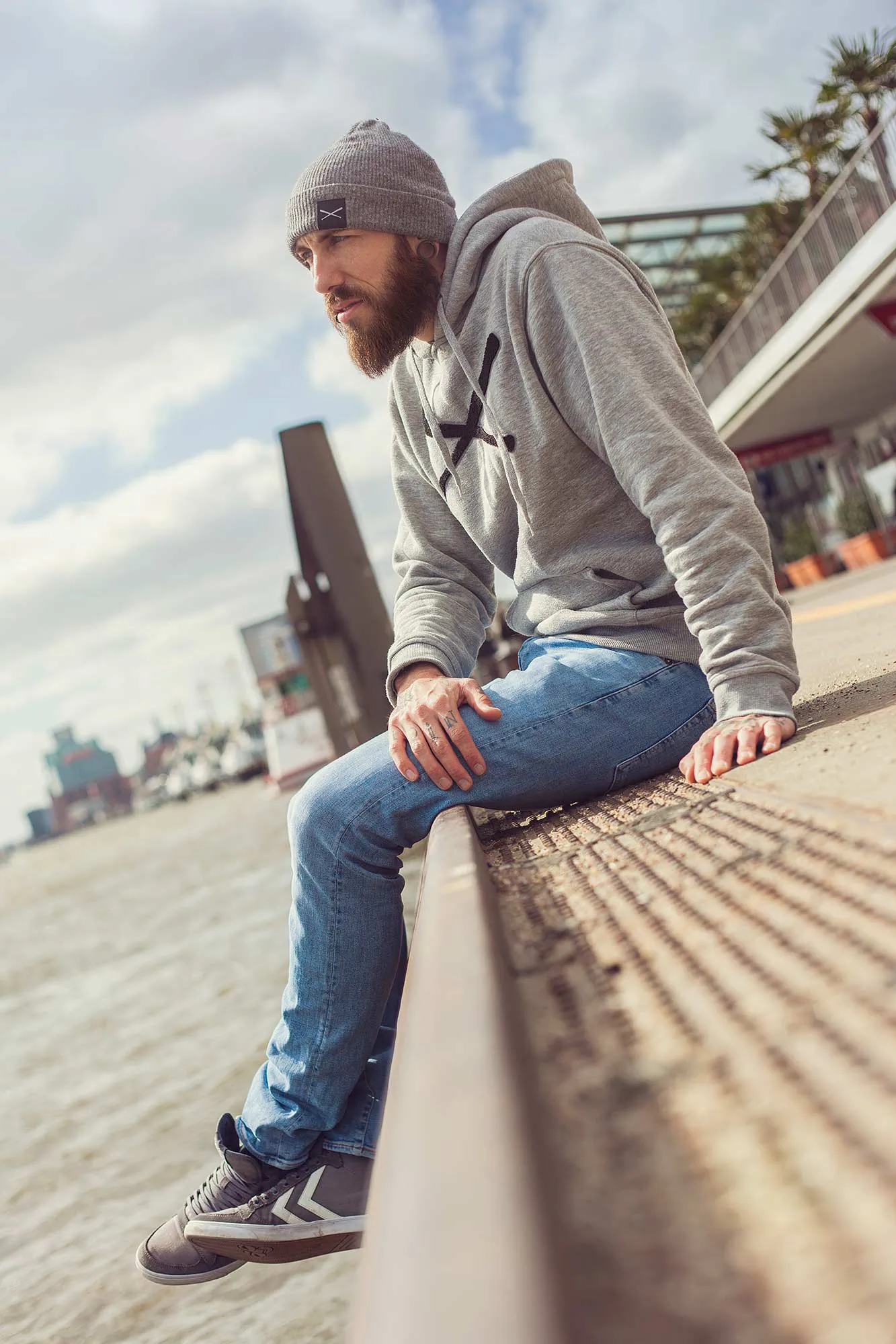 Bärtiger Mann mit grauer Mütze und Hoodie sitzt bei Tageslicht auf einer Uferkante am Wasser.