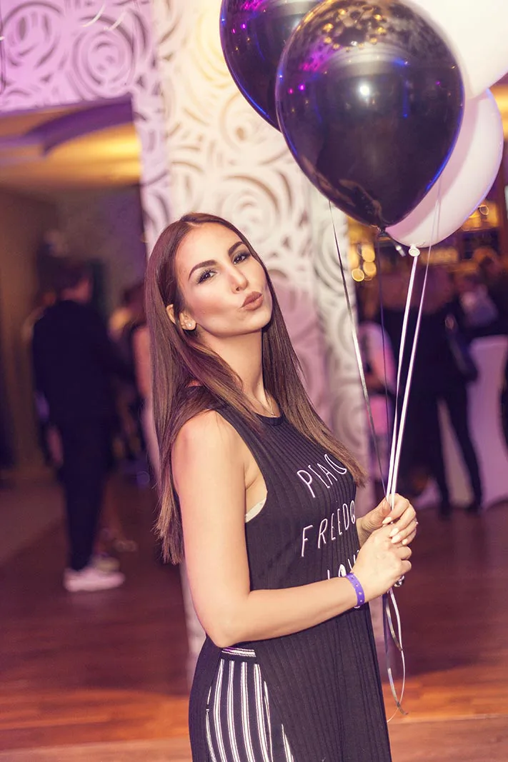 Frau im schwarzen Kleid hält schwarze und weiße Luftballons im violett beleuchteten Eventraum.