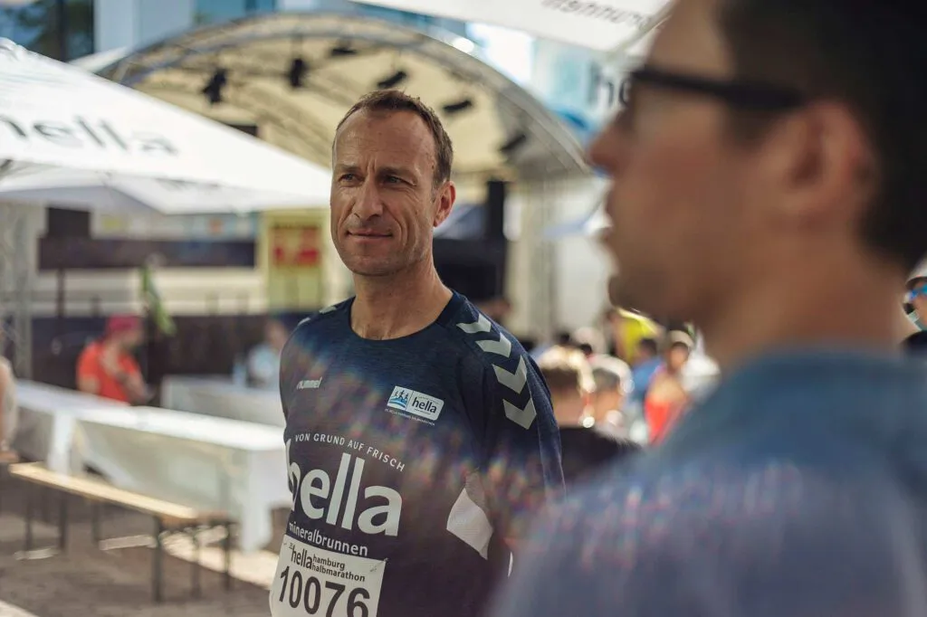 Läufer im hella-Trikot mit Startnummer 10076 im Sonnenlicht, rechts eine unscharfe Person mit Brille.