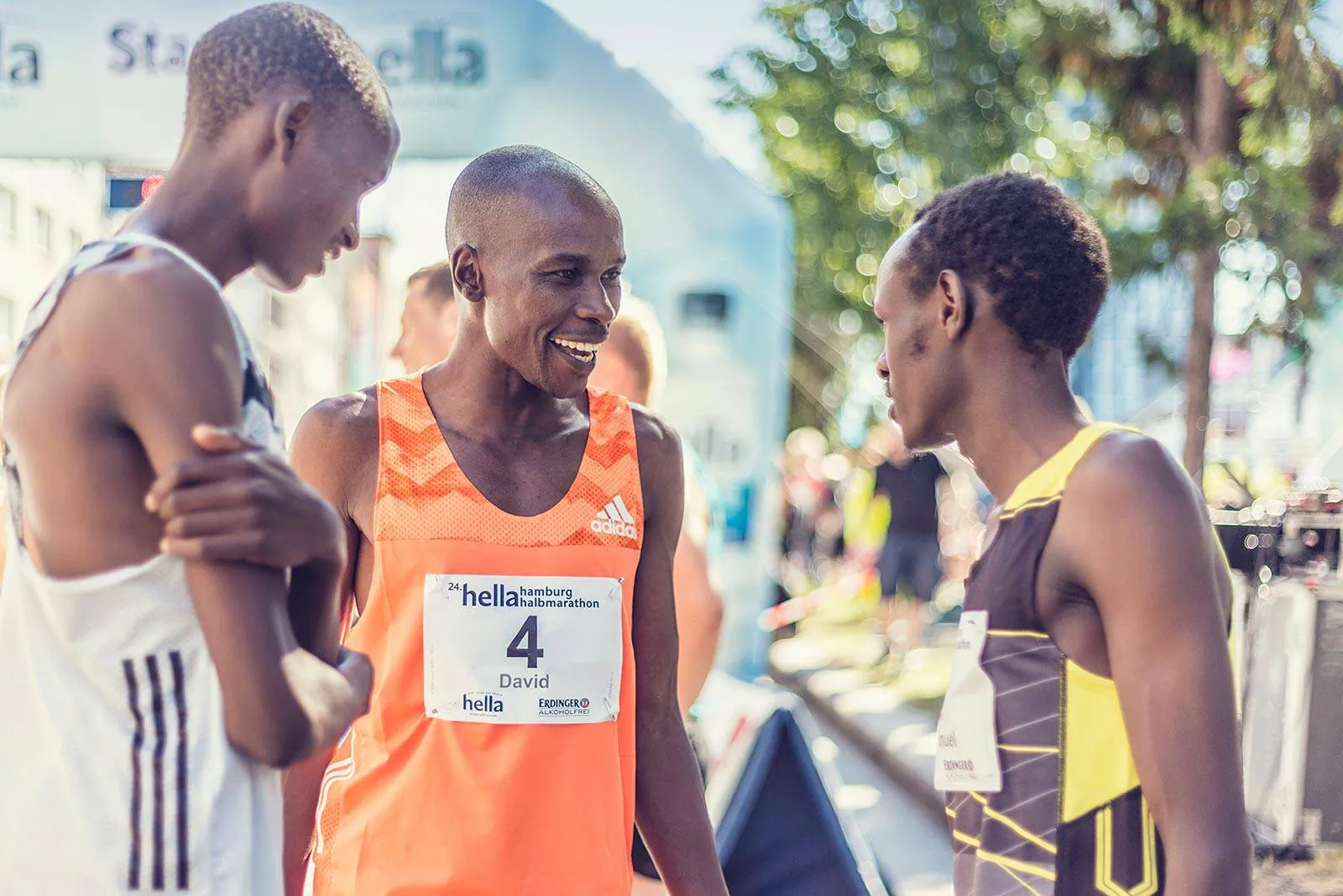 Drei Läufer in Sporttrikots stehen beim Hella Halbmarathon im Sonnenlicht zusammen, einer lächelt.