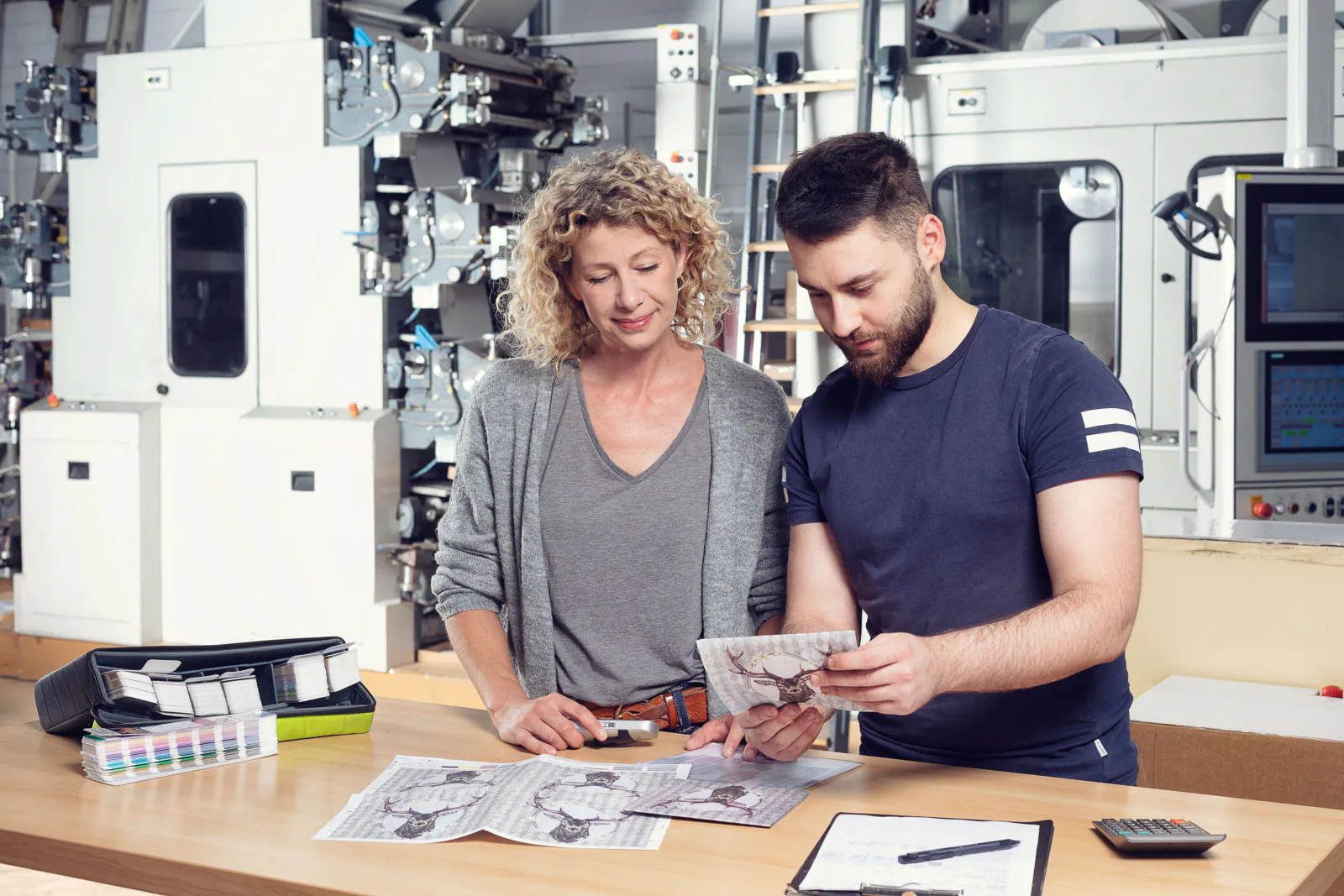 Frau und Mann prüfen Druckmuster an einem Tisch mit Farbfächer vor großen Maschinen in einer hellen Druckerei.