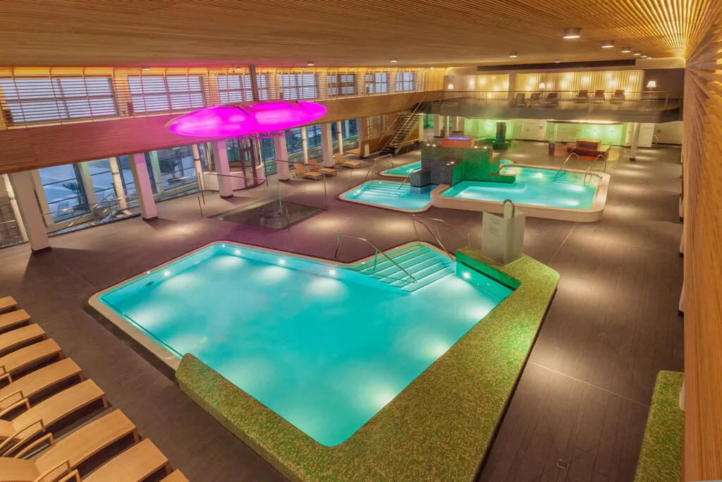 Türkis beleuchtete Innenbecken der VitaSol Therme mit Liegen, Holzdecke und pinker Deckenleuchte, ohne Badegäste.