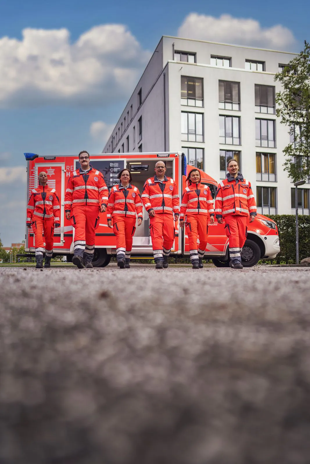 Sechs Johanniter-Rettungskräfte in roten Uniformen vor einem Rettungswagen und modernem Gebäude bei Tageslicht.