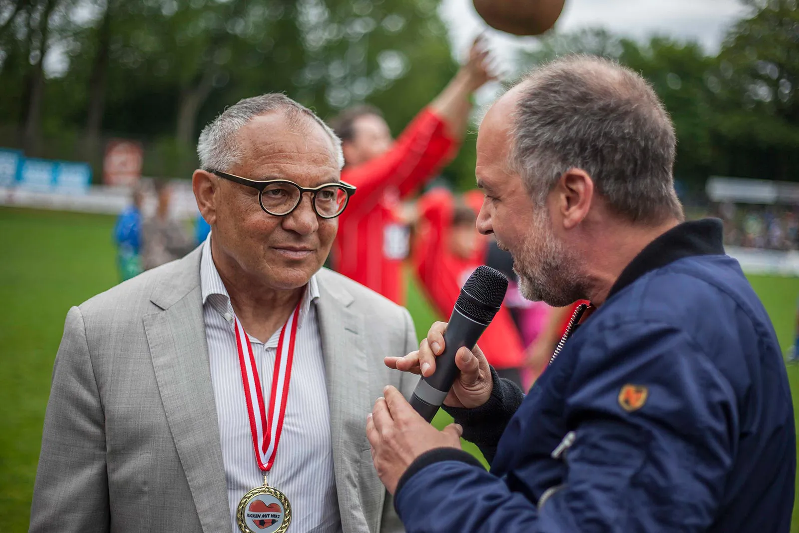 Mann mit Mikrofon interviewt einen Mann mit Brille, grauem Sakko und Medaille am Fußballplatz.