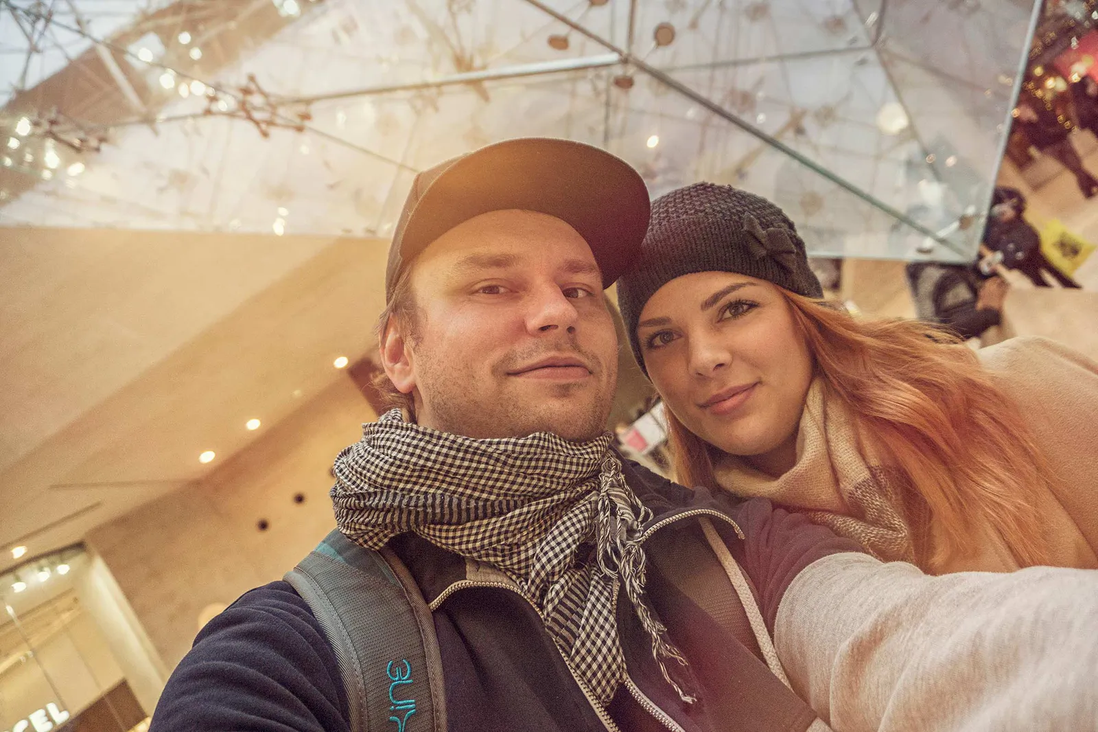 Mann mit Kappe und Schal und Frau mit Strickmütze posieren für ein Selfie unter einem Glasdach im warmen Licht.