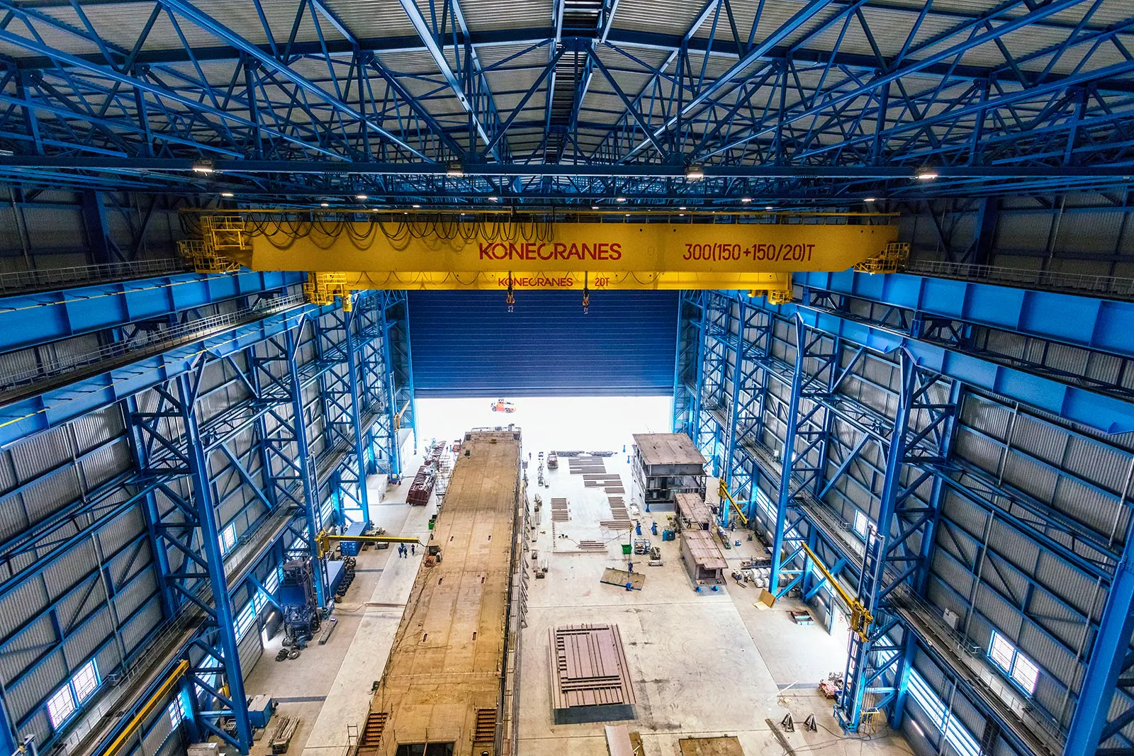 Gelber KONECRANES-Brückenkran in hoher Industriehalle mit blauen Stahlträgern und Tageslicht durch das offene Tor.