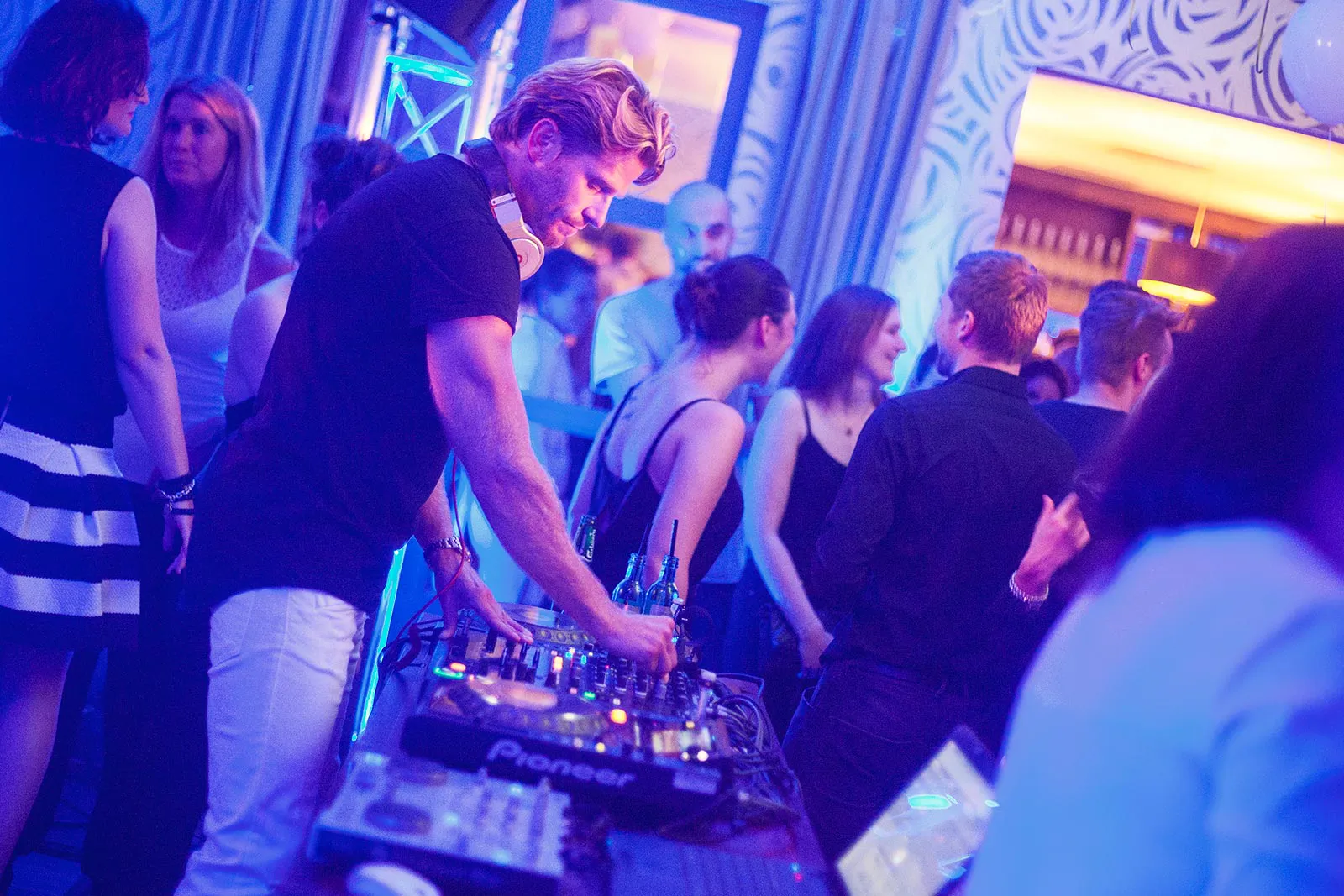 DJ mit Kopfhörern am Mischpult, umgeben von Partygästen im blau-violett beleuchteten Innenraum.