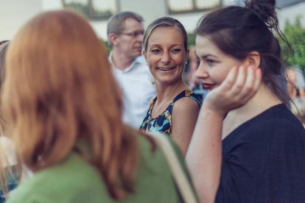 Drei Frauen stehen bei Tageslicht im Freien zusammen, eine lächelt, eine andere stützt das Kinn in die Hand.