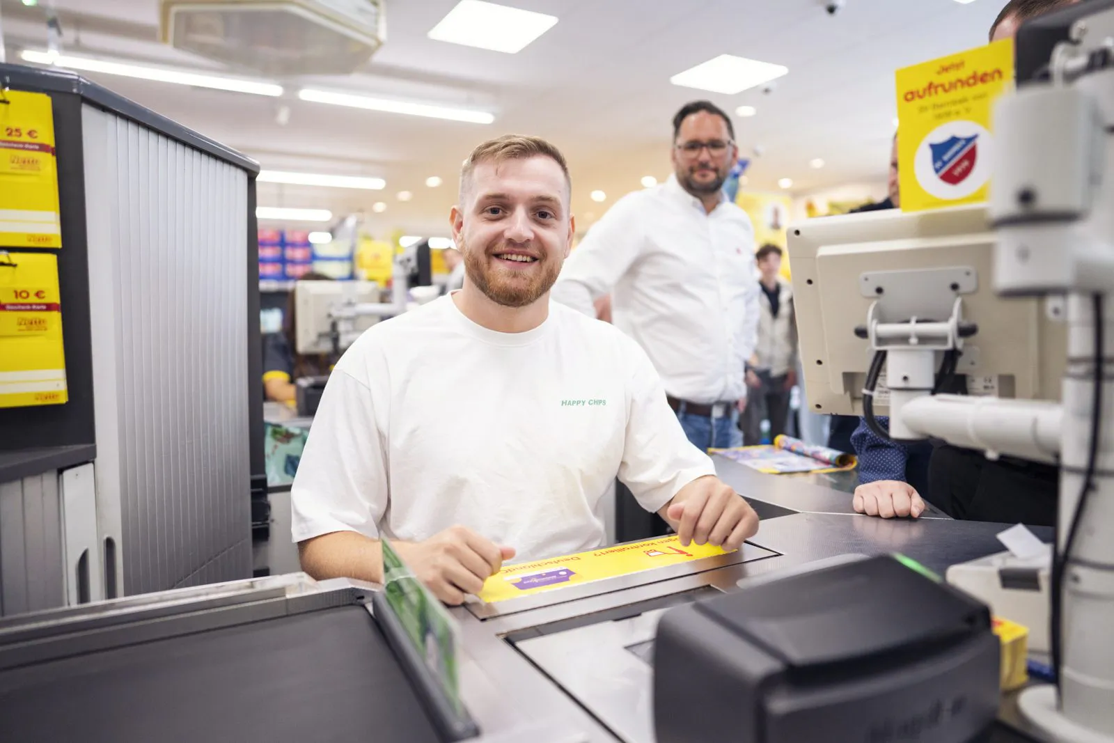Lächelnder Mann im weißen Happy-Chips-Shirt sitzt an einer hell beleuchteten Netto-Kasse, weitere Personen dahinter.