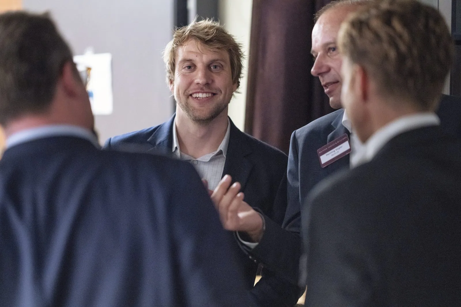Vier Männer in Anzügen unterhalten sich bei einem Event in einem hellen Innenraum, einer lächelt.