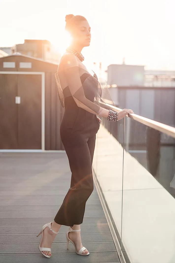 Frau in schwarzem Outfit und weißen High Heels am Glasgeländer einer Dachterrasse im warmen Gegenlicht.