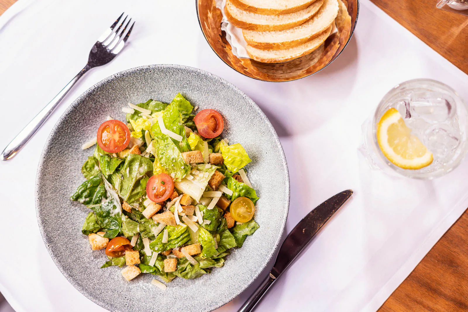 Salat mit Tomaten und weiteren Zutaten auf einem grauen Teller, daneben Brot und ein Glas mit Zitronenscheibe auf weiß gedecktem Tisch.