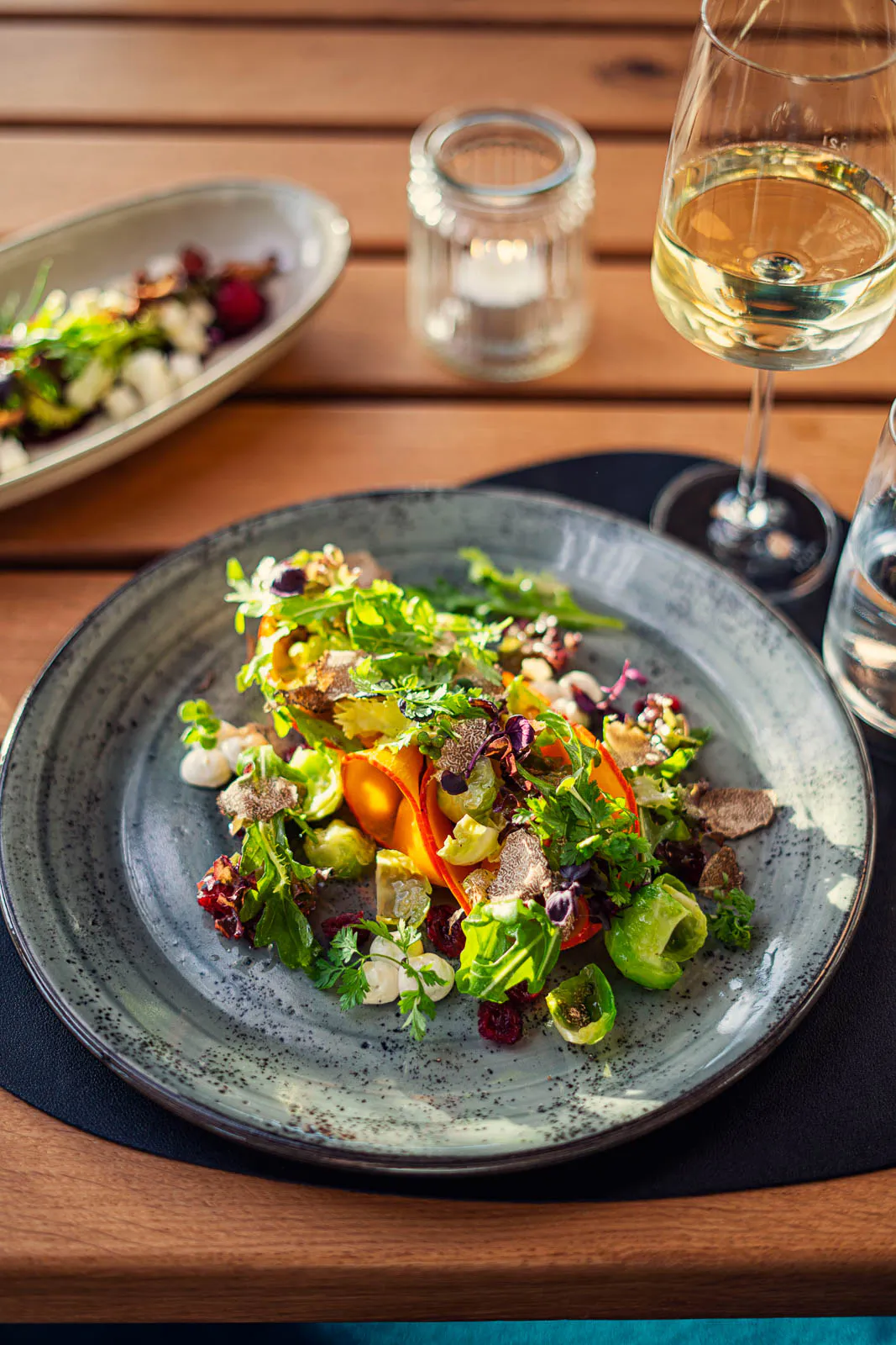 Bunter Salat mit Kräutern und Gemüse auf grauem Teller, daneben Weißwein und Windlicht auf sonnigem Holztisch.