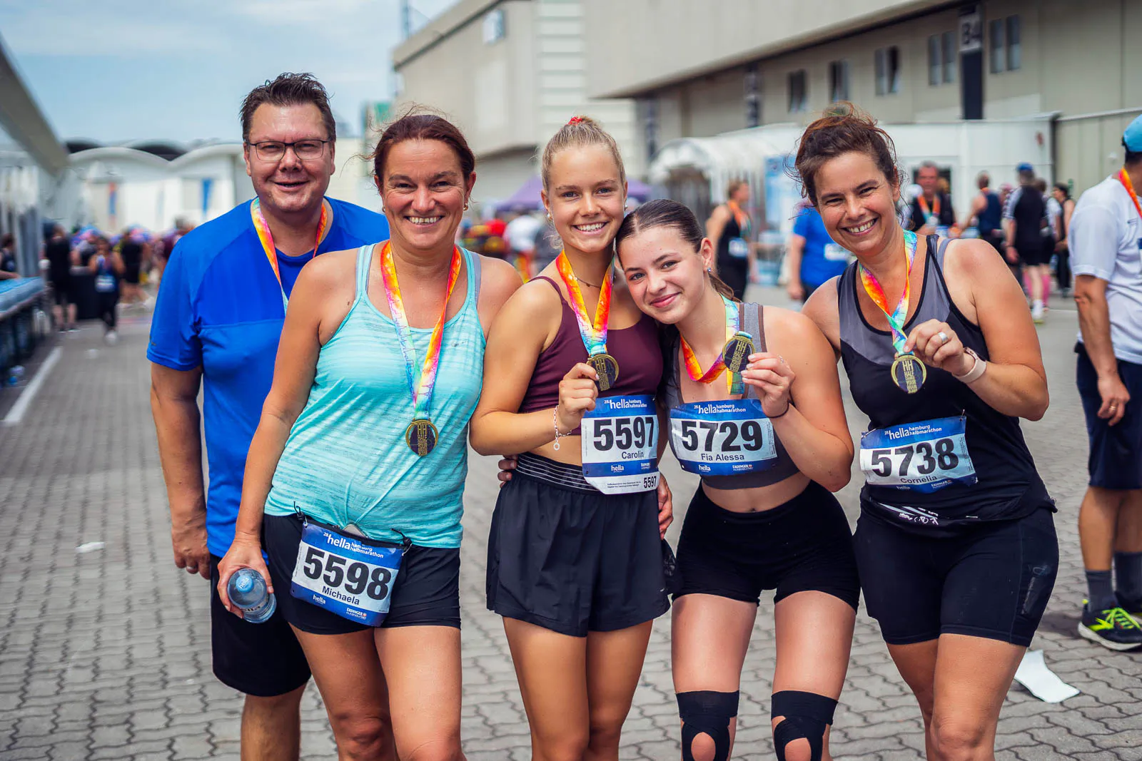 Fünf lächelnde Teilnehmende mit Medaillen posieren beim Hella Halbmarathon vor einem Gebäude im Tageslicht.