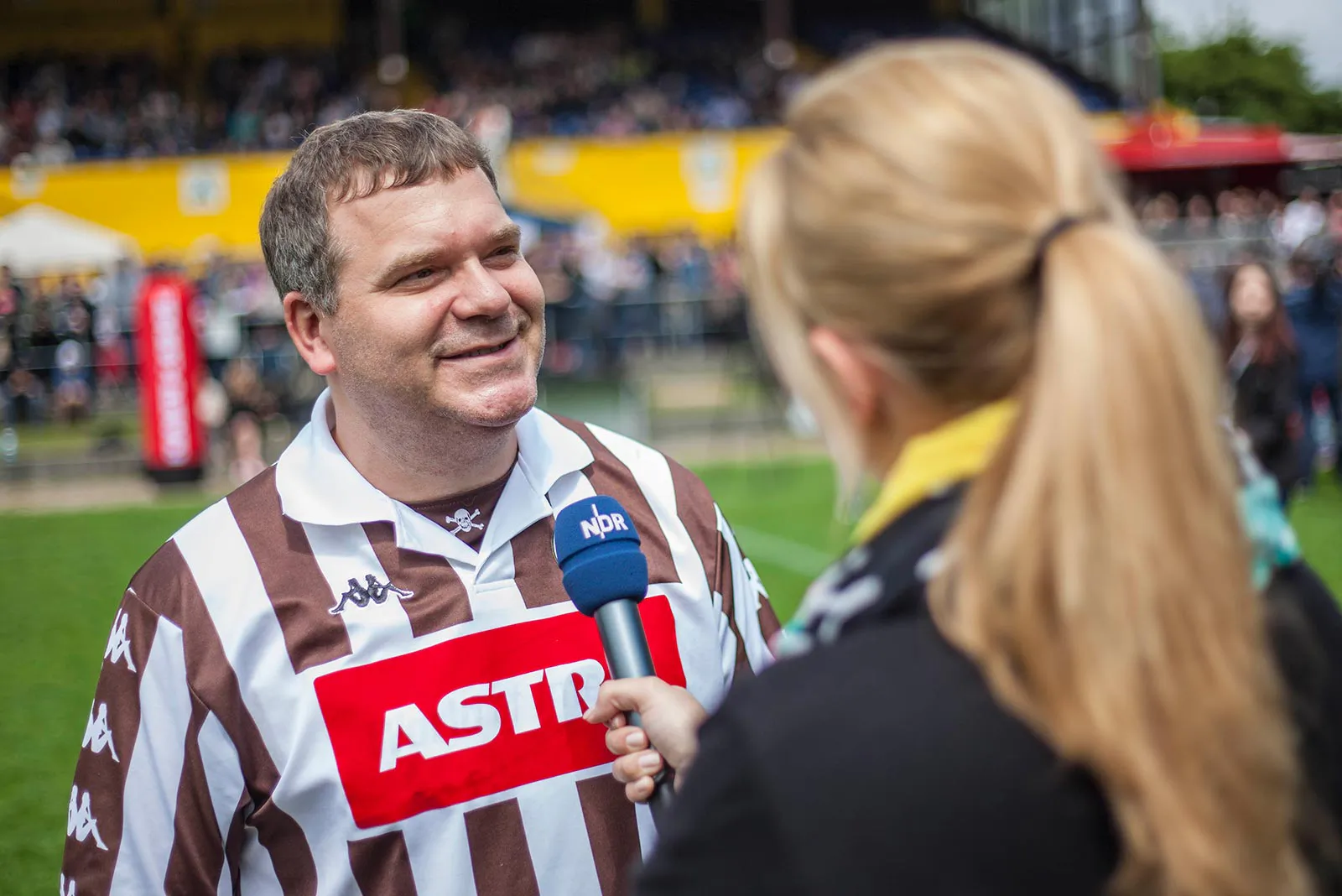 Ein Mann im braun-weiß gestreiften Fußballtrikot wird am Spielfeldrand von einer Frau mit Mikrofon interviewt.