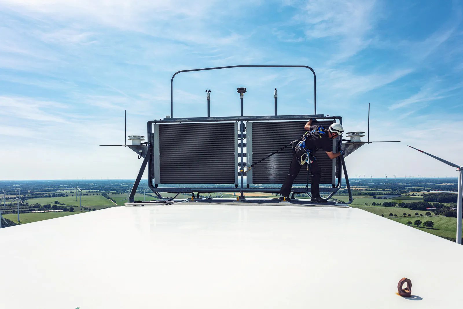 Gesicherter Techniker mit Helm arbeitet auf einer Windkraftanlage, im Hintergrund Felder und Windräder bei Tageslicht.