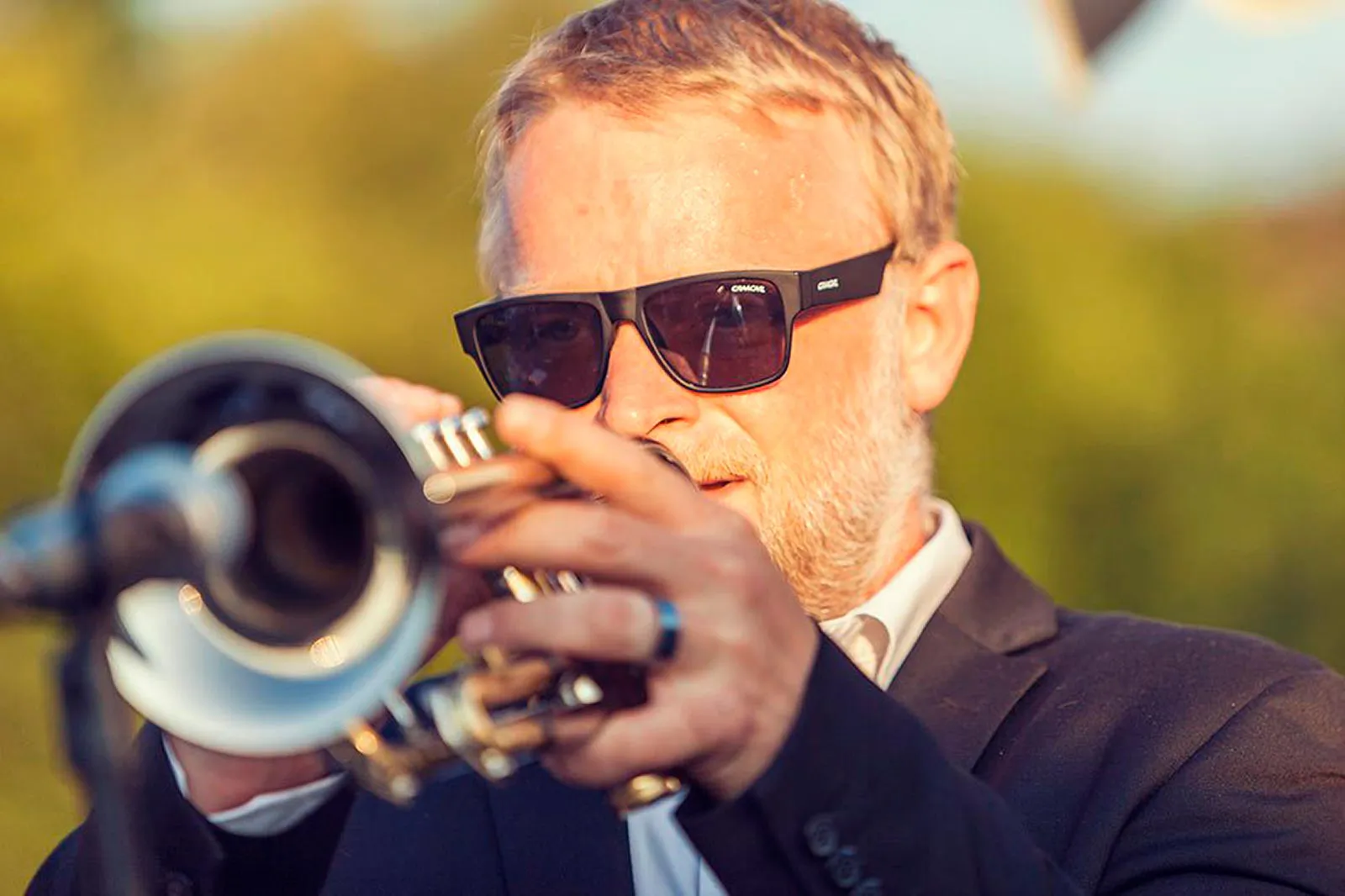 Mann mit Sonnenbrille und dunklem Sakko spielt Trompete vor einem Mikrofon im warmen Sonnenlicht im Freien.