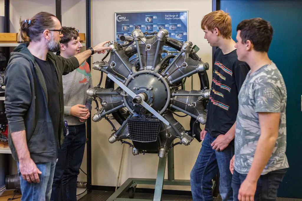 Ein Mann erklärt drei Jugendlichen einen großen Sternmotor in einem technischen Unterrichtsraum.