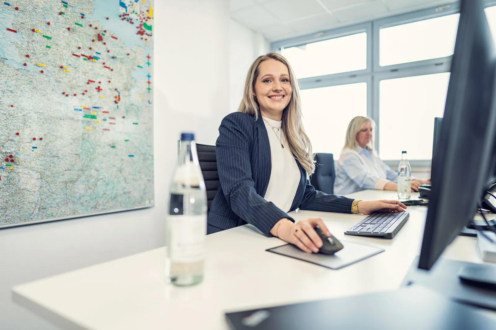 Zwei Frauen an Computern im hellen Büro, Mineralwasserflaschen auf den Tischen und eine große Karte an der Wand.