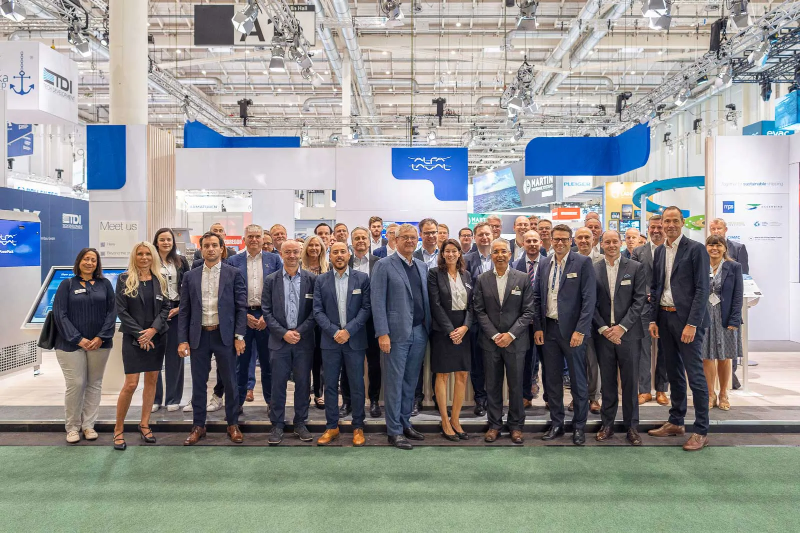 Große Gruppe in Geschäftskleidung posiert am hell beleuchteten Messestand von Alfa Laval in einer Messehalle.