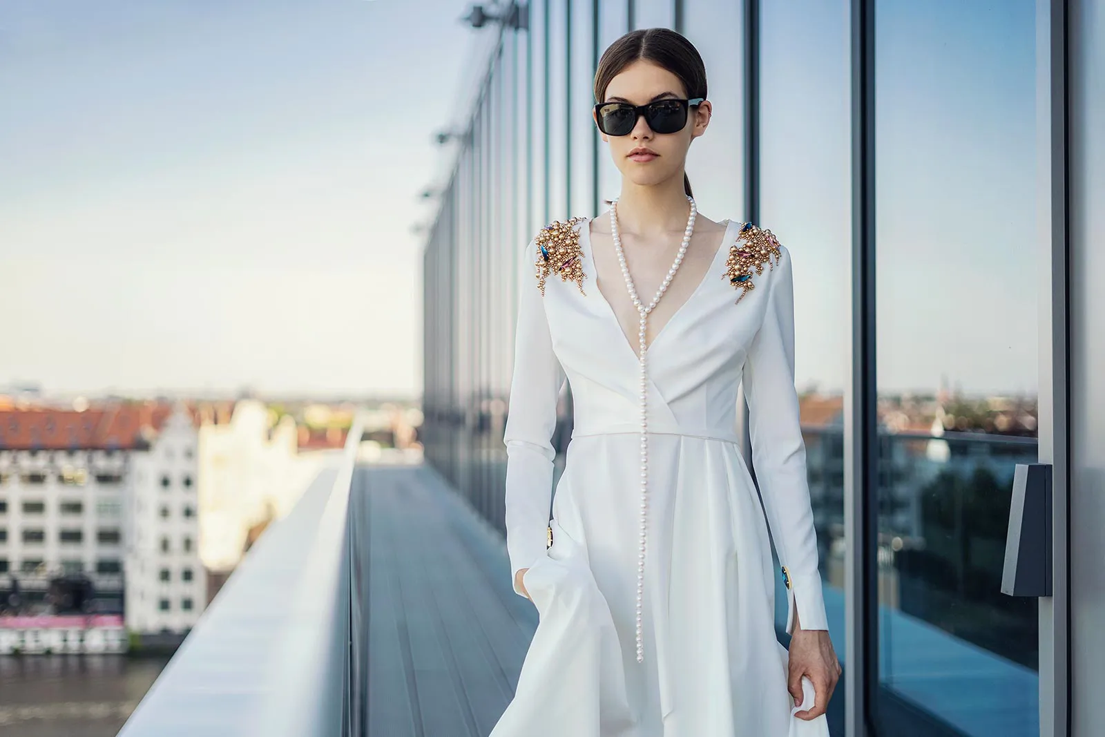 Frau in weißem Kleid mit Schulterverzierungen, Perlenkette und Sonnenbrille auf einer Terrasse vor einer Glasfassade.