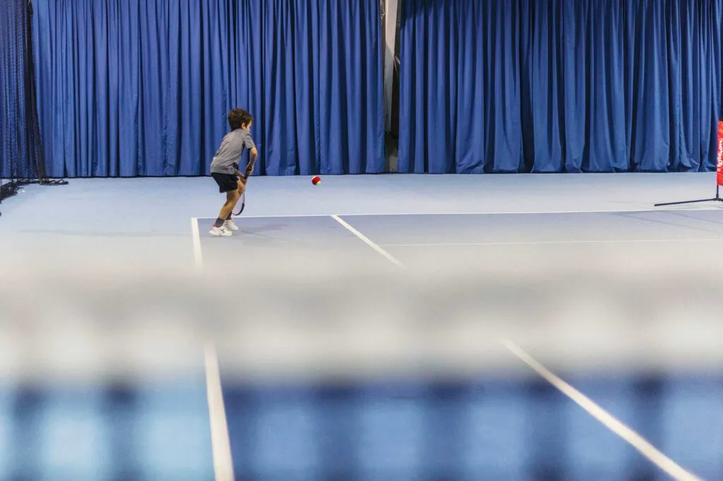 Junger Tennisspieler mit Schläger läuft zum Ball auf blauem Hallenplatz, unscharfes Netz im Vordergrund.