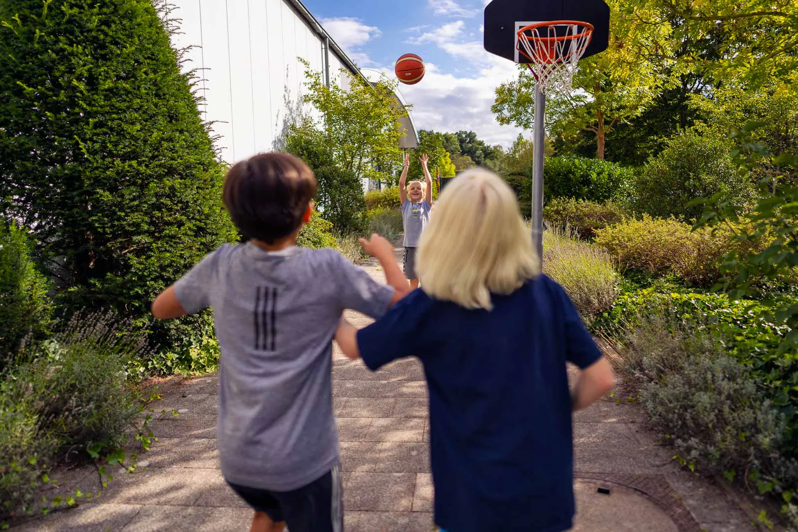 Drei Kinder spielen bei Tageslicht Basketball auf einem gepflasterten Weg zwischen Hecken und einem Gebäude.