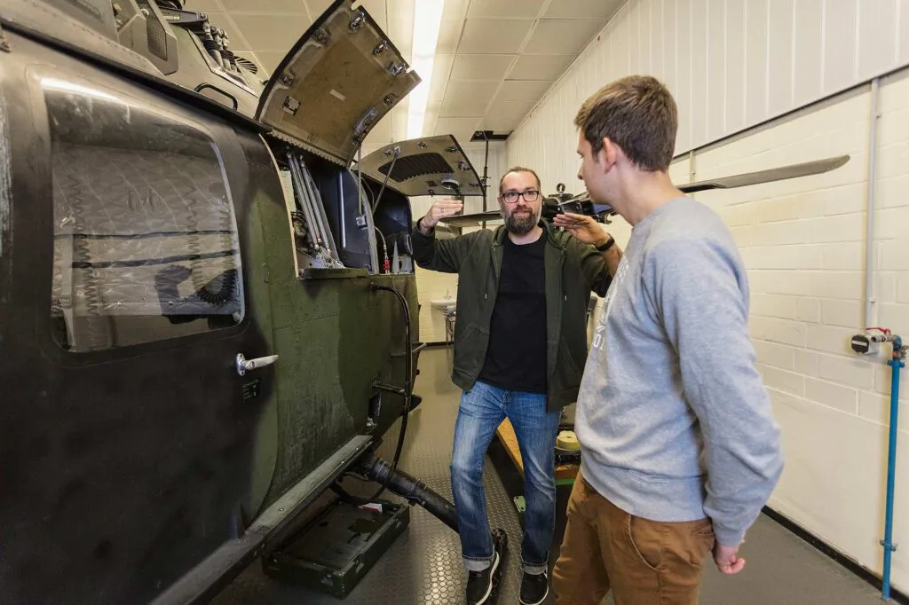 Zwei Männer stehen neben einem geöffneten Hubschrauber in einer hellen Werkhalle; einer gestikuliert erklärend.