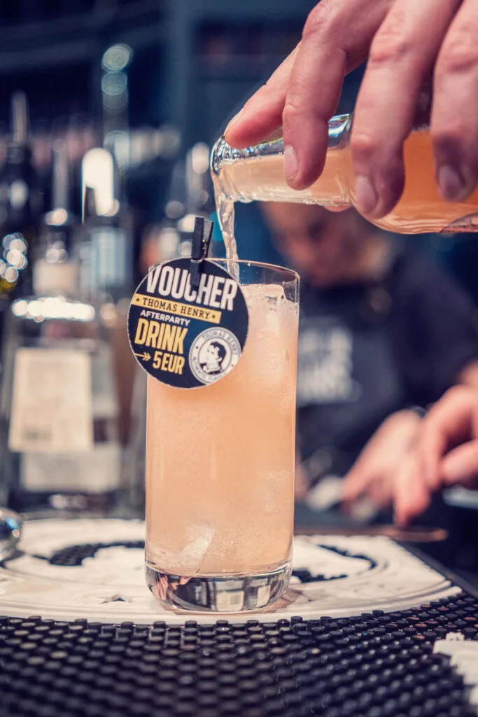 Eine Person gießt an einer Bar ein orangefarbenes Getränk in ein Glas mit Eis und Thomas-Henry-Gutschein.