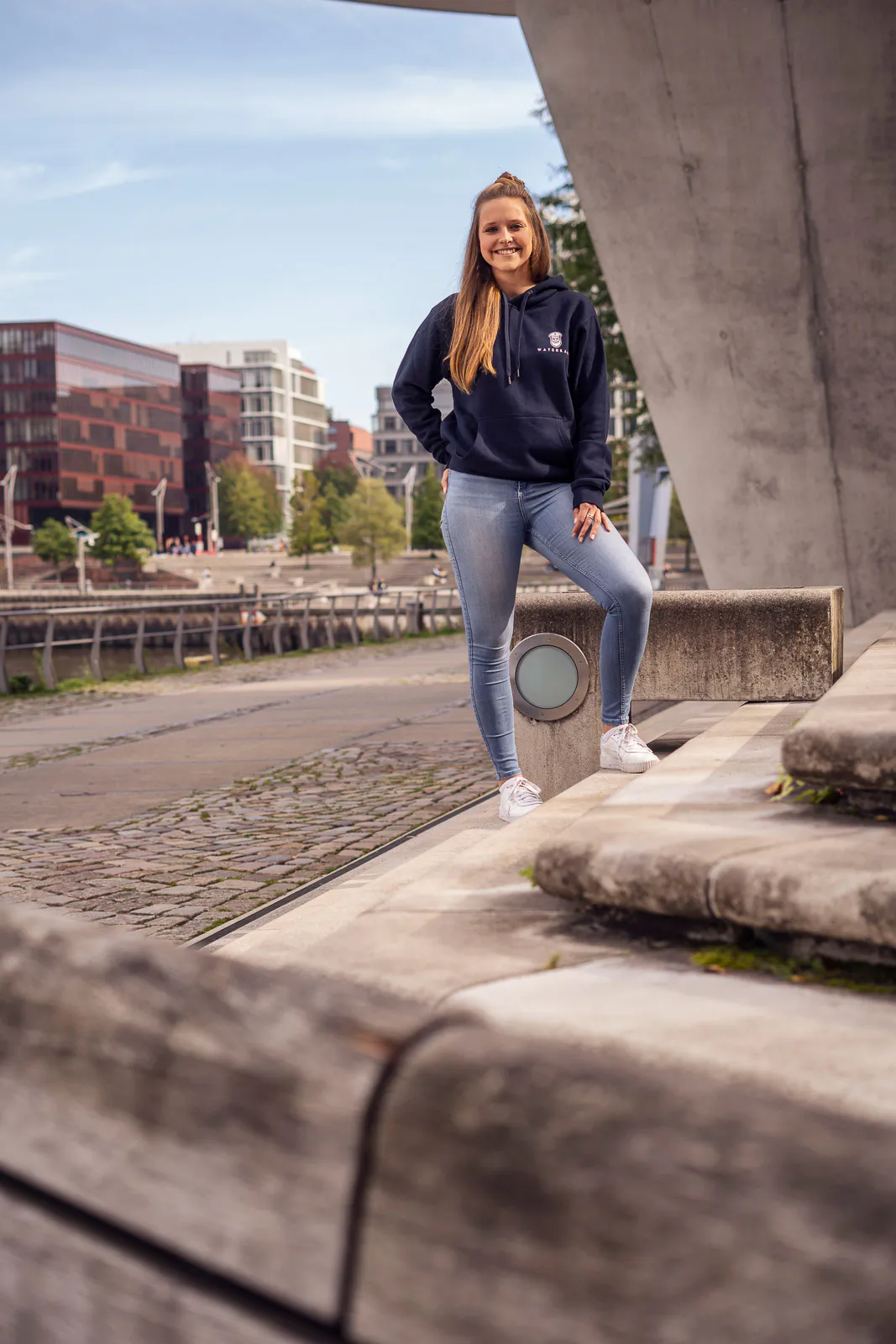 Frau im dunklen Hoodie und Jeans steht lächelnd auf sonnigen Stufen am Wasser neben einer Betonkonstruktion.