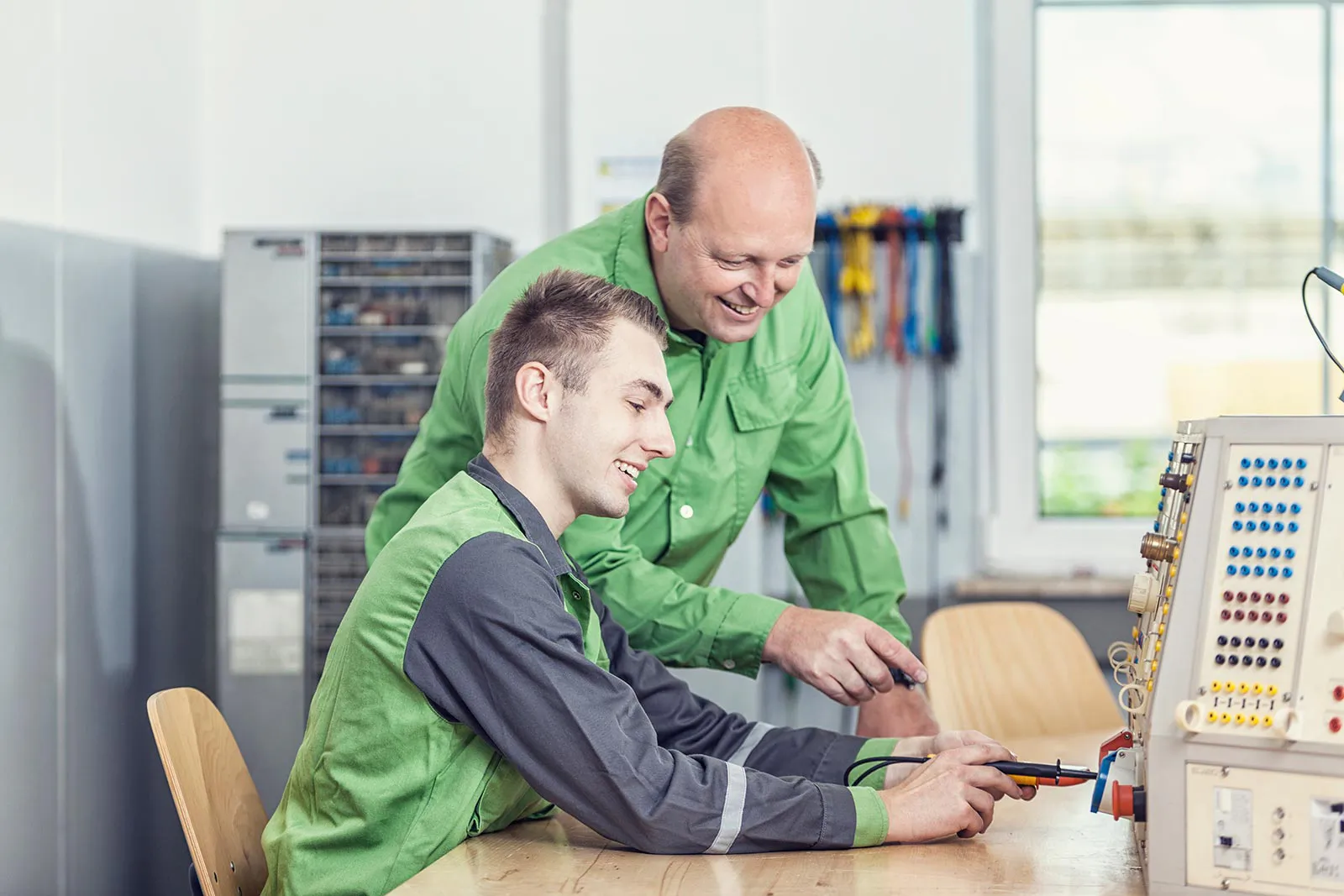 Zwei Männer in grüner Arbeitskleidung arbeiten in einer hellen Werkstatt an einem elektrischen Übungsgerät.