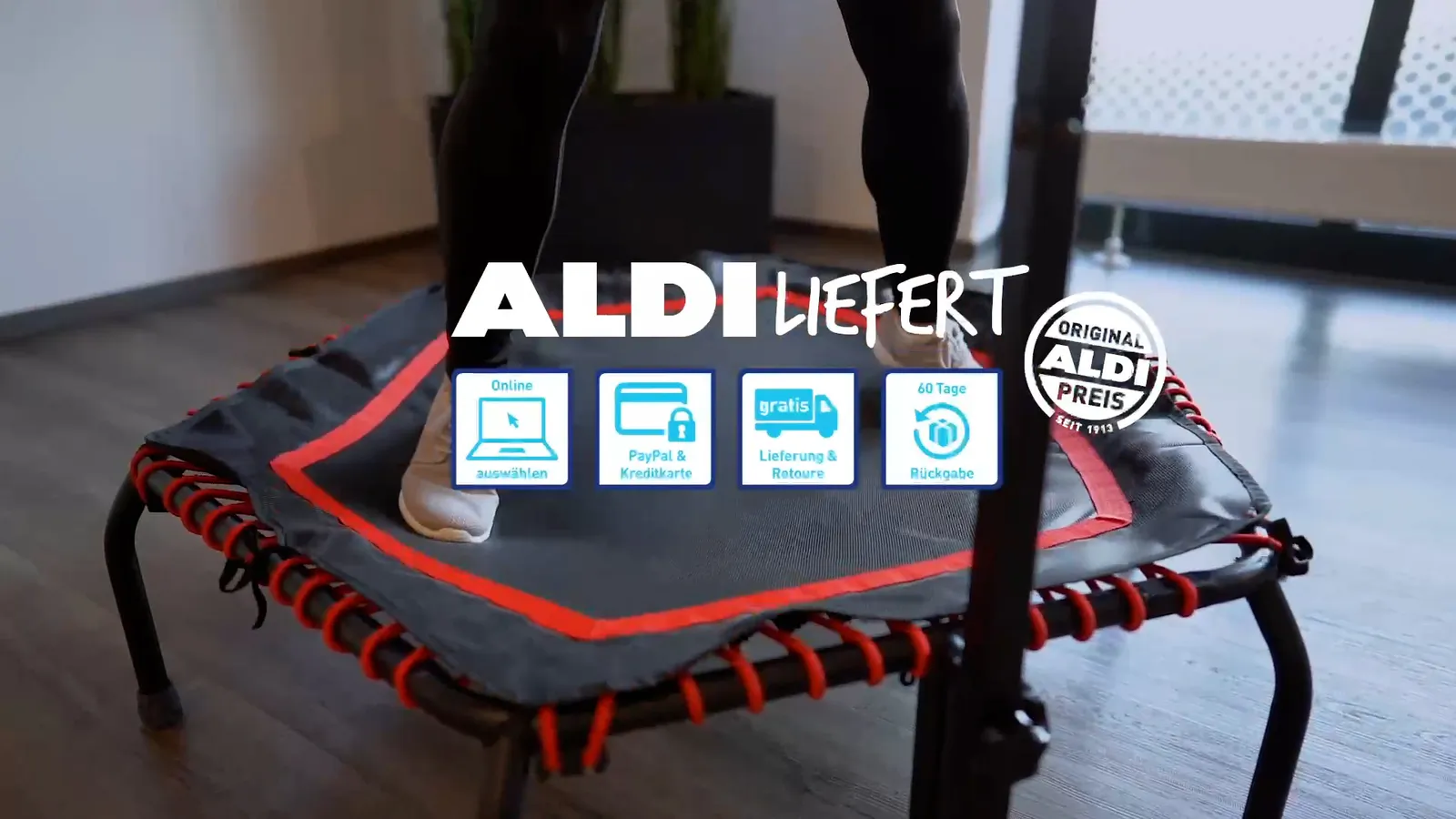 Rot-schwarzes Minitrampolin mit Haltegriff in einem hellen Wohnraum, darüber der Schriftzug ALDI LIEFERT und Markenlogos.