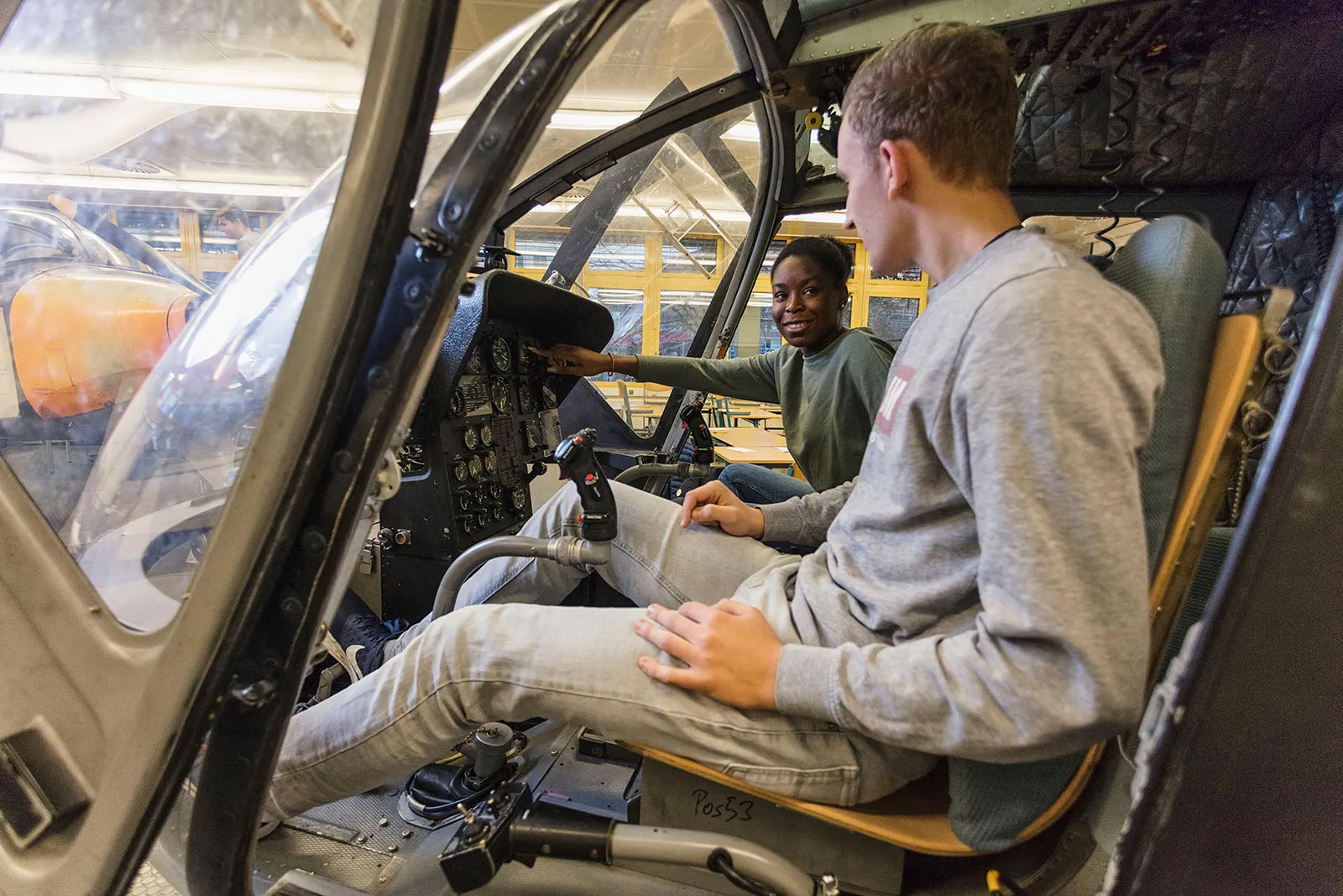 Zwei junge Männer sitzen im Hubschraubercockpit in einer hellen Werkhalle; einer zeigt auf die Instrumente.