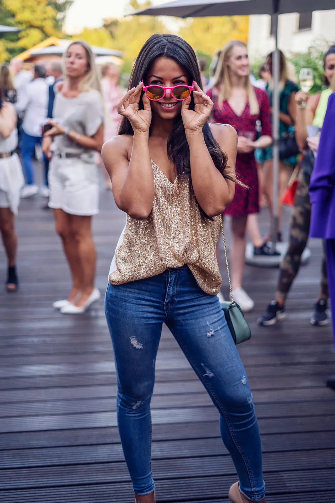 Lächelnde Frau mit pinker Sonnenbrille, Glitzertop und Jeans auf einer Holzterrasse mit Partygästen im Tageslicht.
