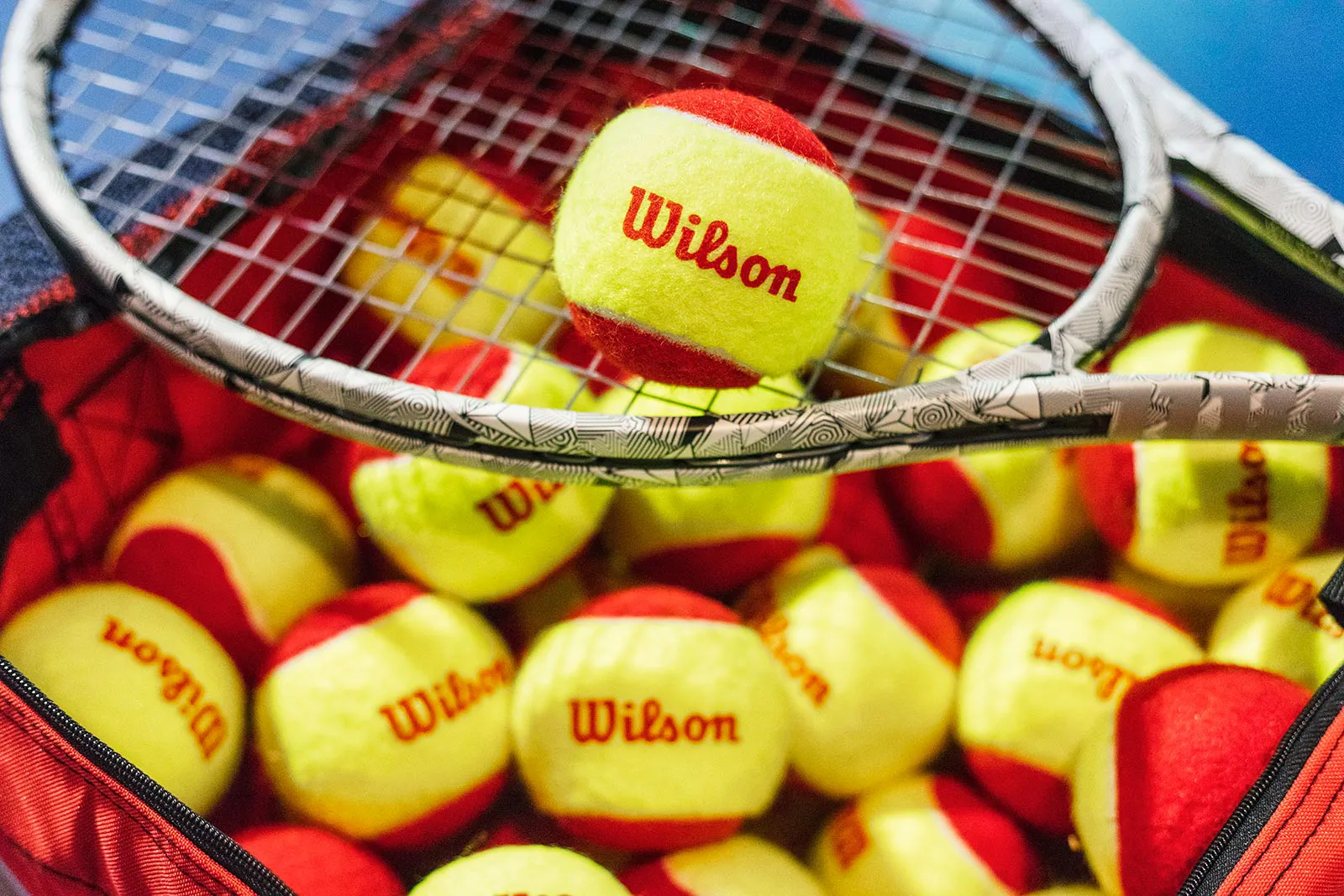 Gelb-rote Wilson-Tennisbälle in einer offenen Tasche, darüber ein Tennisschläger mit einem Ball auf der Bespannung.