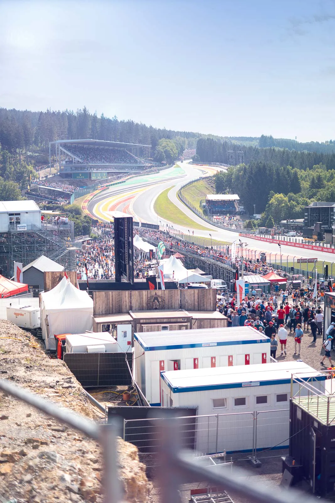 Sonnige Formel-1-Rennstrecke mit steiler Kurvenpassage, Tribünen und vielen Zuschauern zwischen Zelten und Containern.