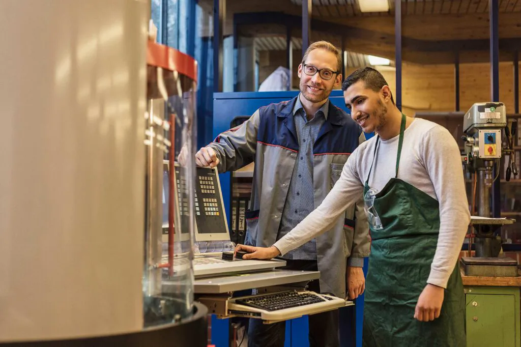 Zwei Männer bedienen eine Maschine in einer Werkstatt, einer trägt eine grüne Schürze, der andere einen Arbeitskittel.