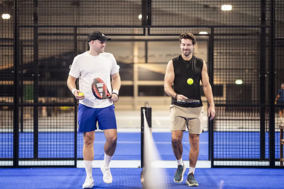Zwei Männer mit Padelschlägern am Netz eines blauen Hallenplatzes; einer lässt einen gelben Ball über dem Schläger springen.