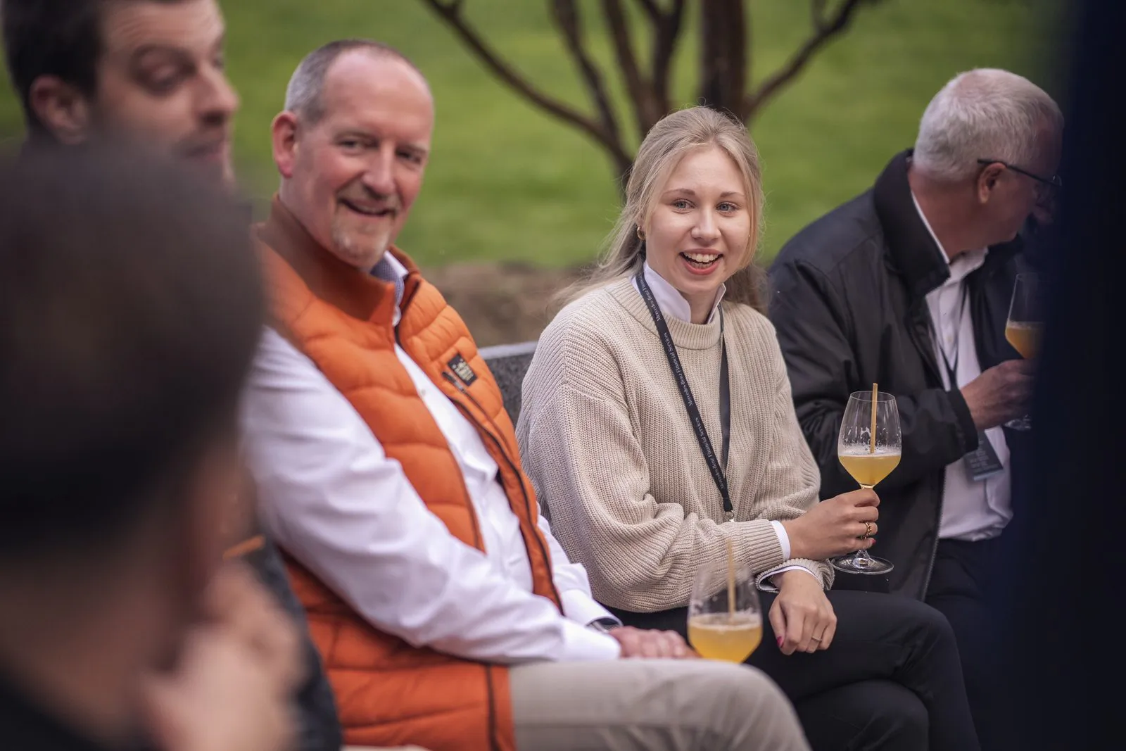 Gäste sitzen bei Tageslicht im Freien, eine lächelnde Frau hält ein Glas mit orangefarbenem Getränk.
