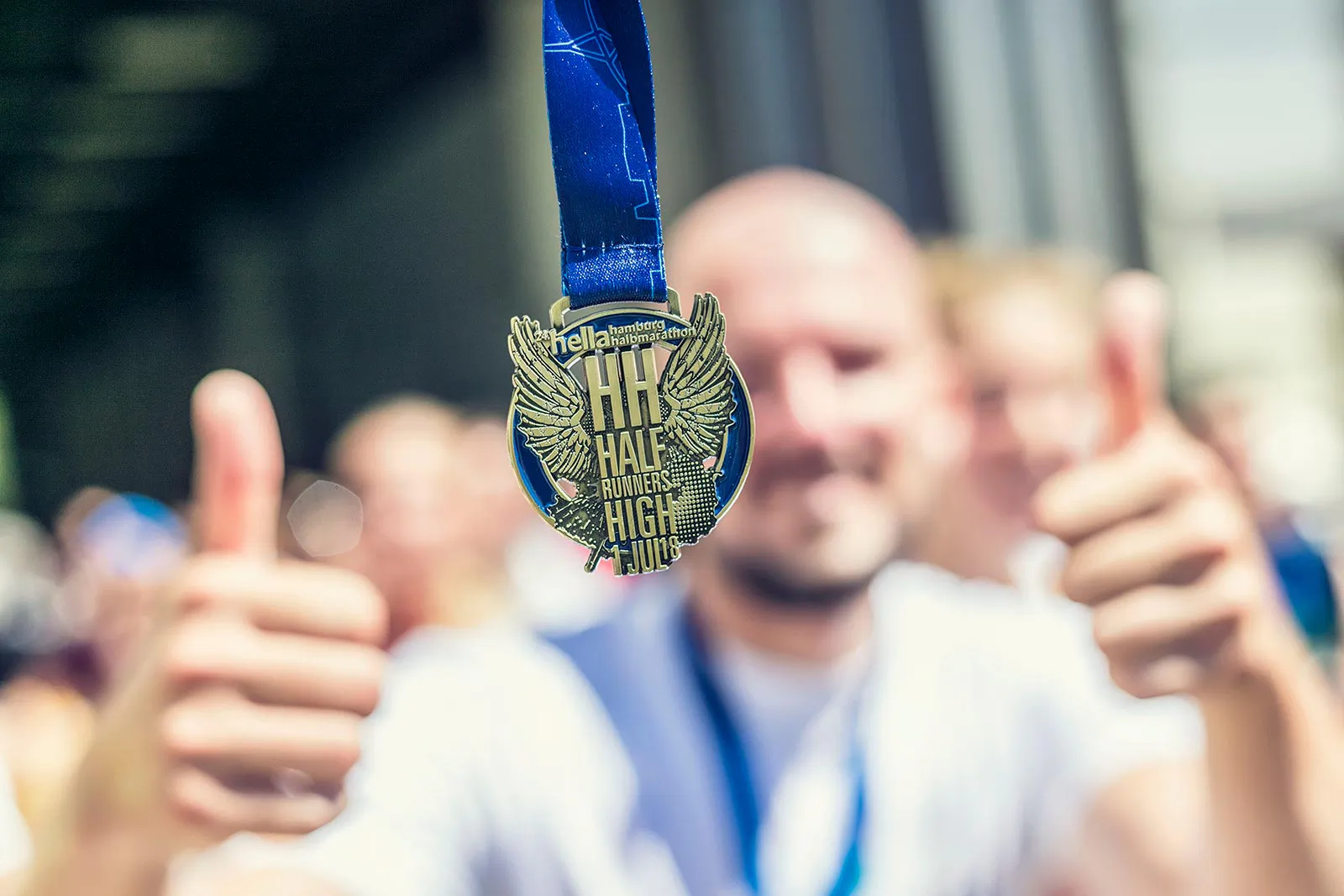 Medaille des Hella Halbmarathons am blauen Band, dahinter ein unscharfer Mann mit beiden Daumen nach oben im Sonnenlicht.