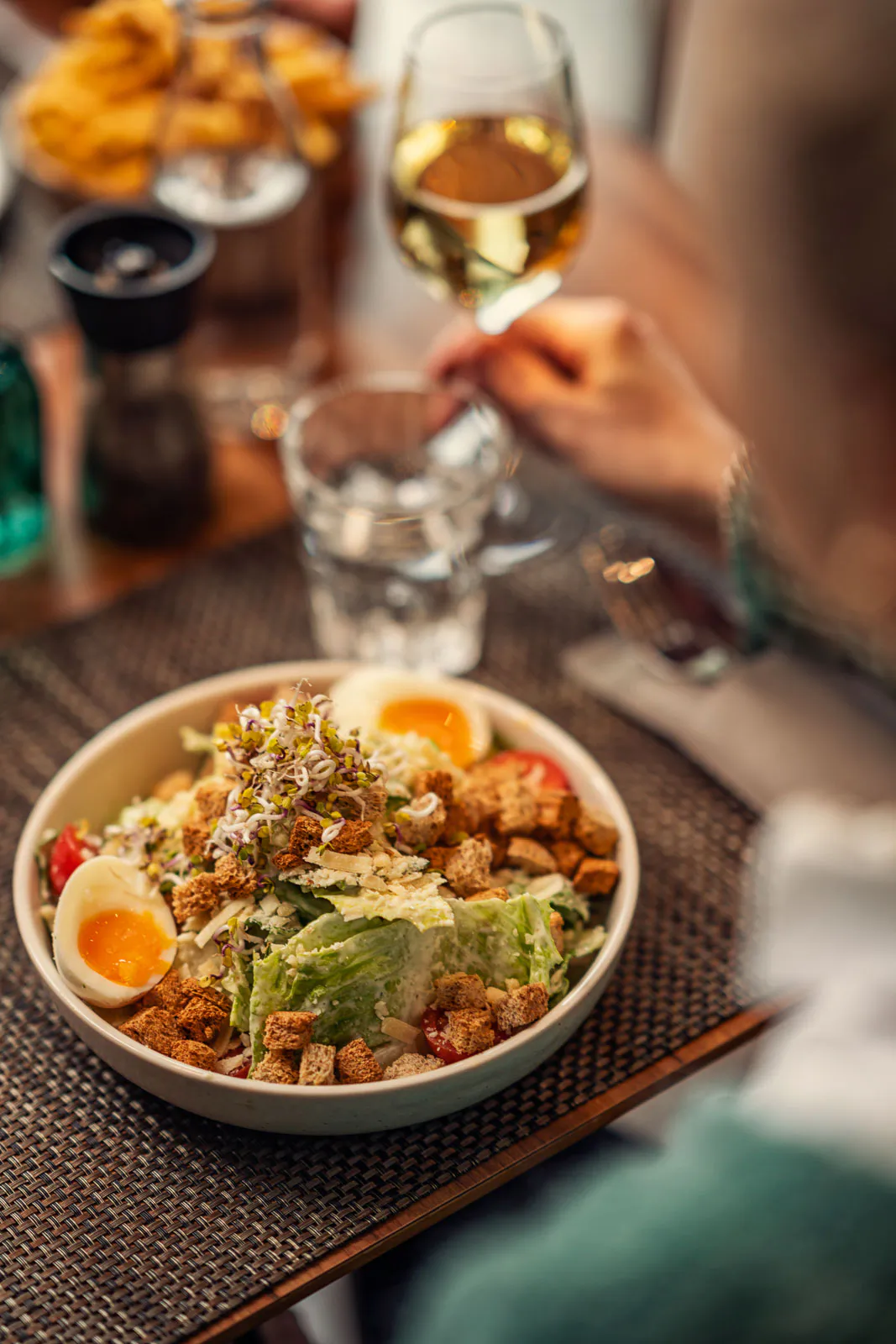 Salatschale mit Ei, Croûtons und Sprossen auf einem Restauranttisch; eine Person hält ein Glas Weißwein.
