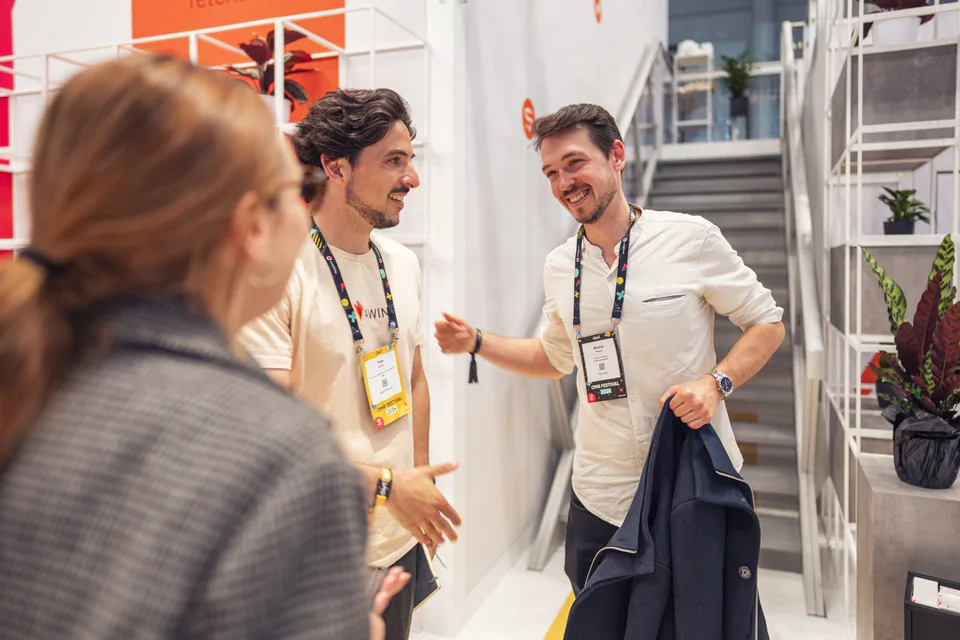 Zwei Männer mit Messeausweisen lachen am hellen Awin-Messestand, eine Frau steht unscharf im Vordergrund.