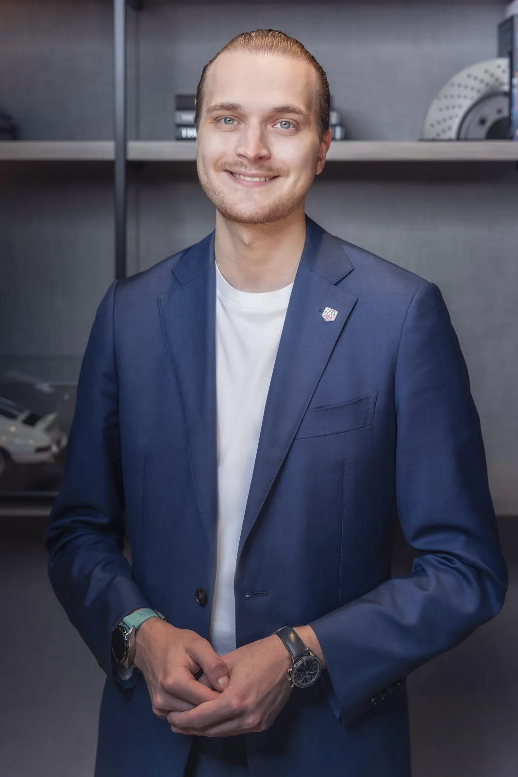 Lächelnder Tag-Heuer-Mitarbeiter im blauen Sakko mit zwei Armbanduhren vor dunklem Regal, weich beleuchtet.