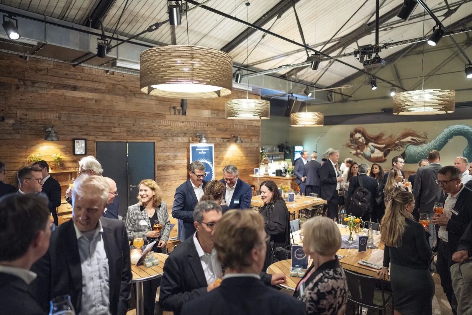 Gäste unterhalten sich an Stehtischen in einer rustikalen Eventlocation mit Holzwänden und warmen Hängelampen.