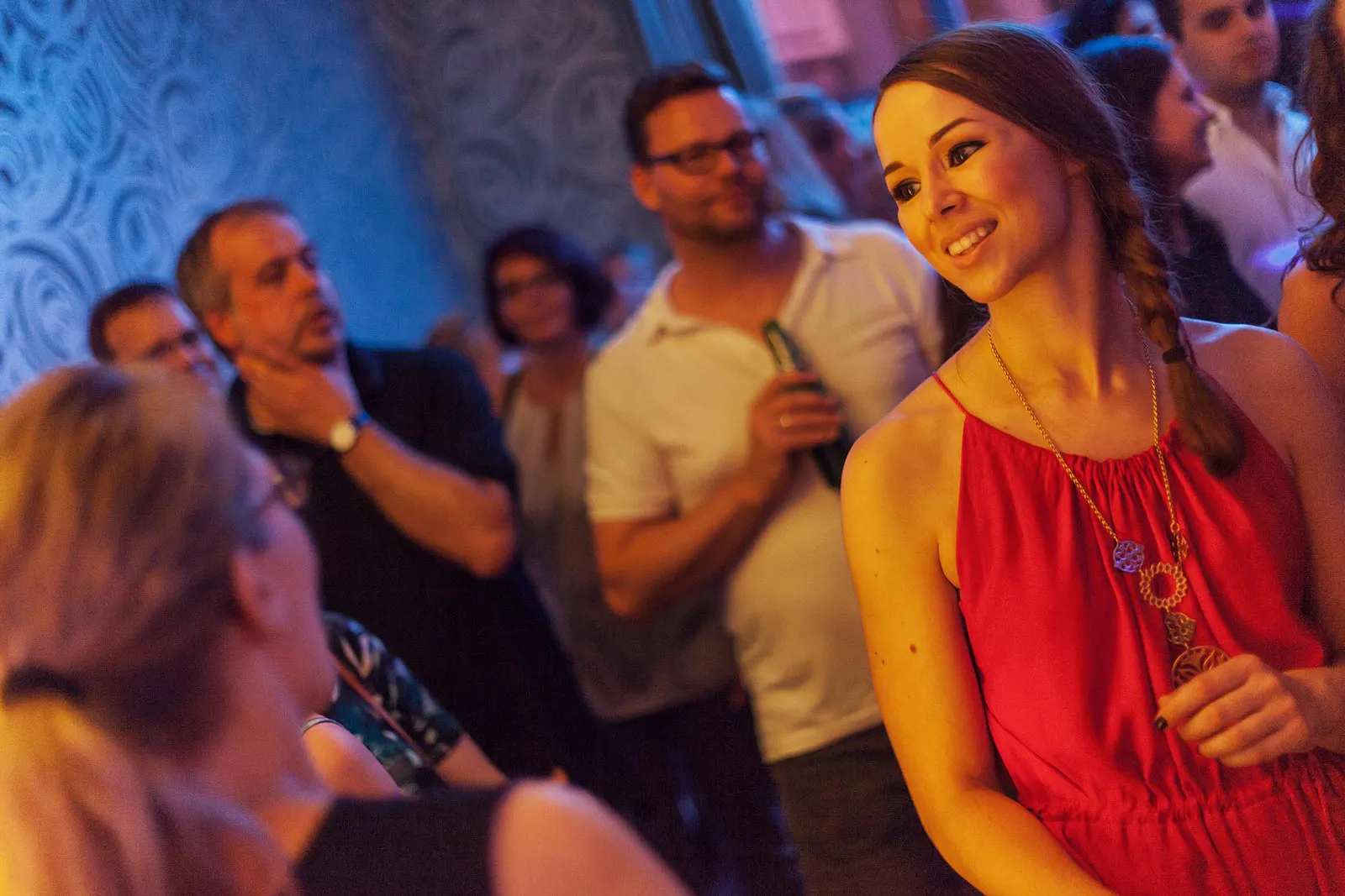 Lächelnde Frau im roten Kleid zwischen Partygästen in einem Innenraum mit blauem und warmem Licht.