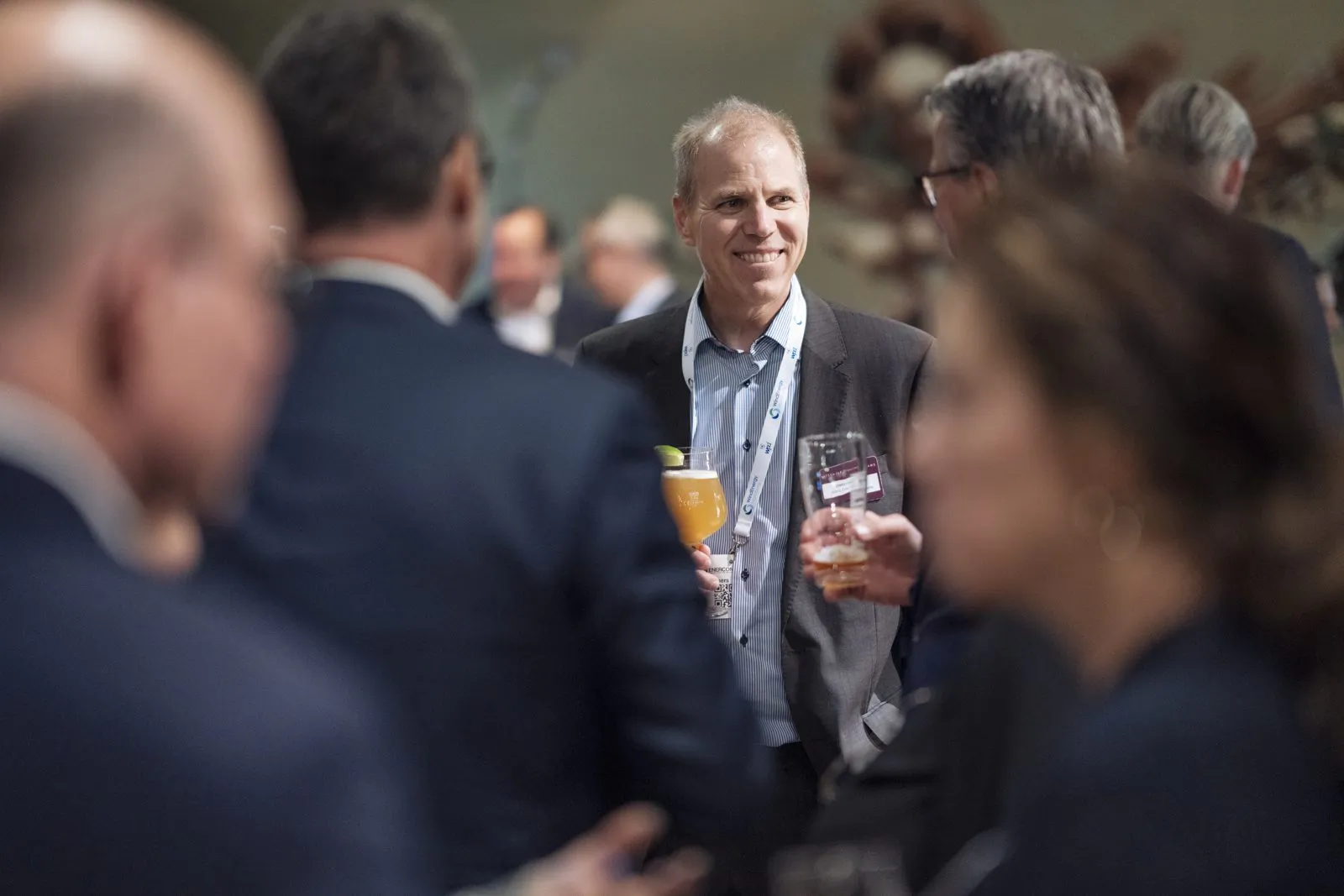 Lächelnder Mann mit orangefarbenem Getränk im Gespräch mit Gästen beim Watson & Farley-Event in einem Innenraum.