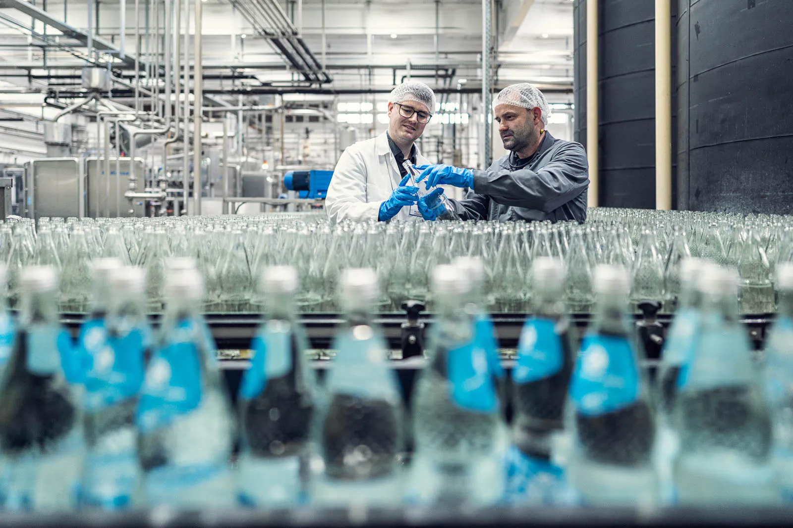 Zwei Mitarbeiter mit Haarnetzen und blauen Handschuhen prüfen Glasflaschen in der hellen Abfüllhalle.