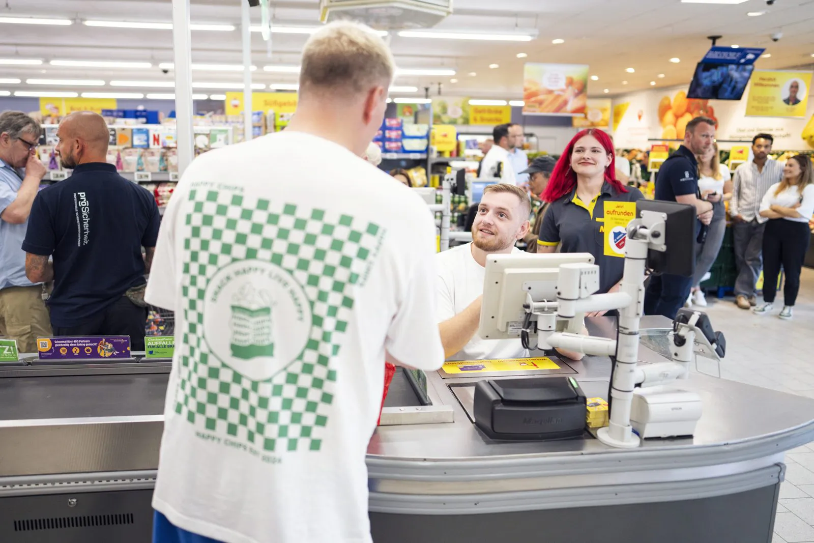 Ein Mann im weißen Happy-Chips-Shirt steht an einer Supermarktkasse, dahinter Personal und Gäste unter hellem Deckenlicht.