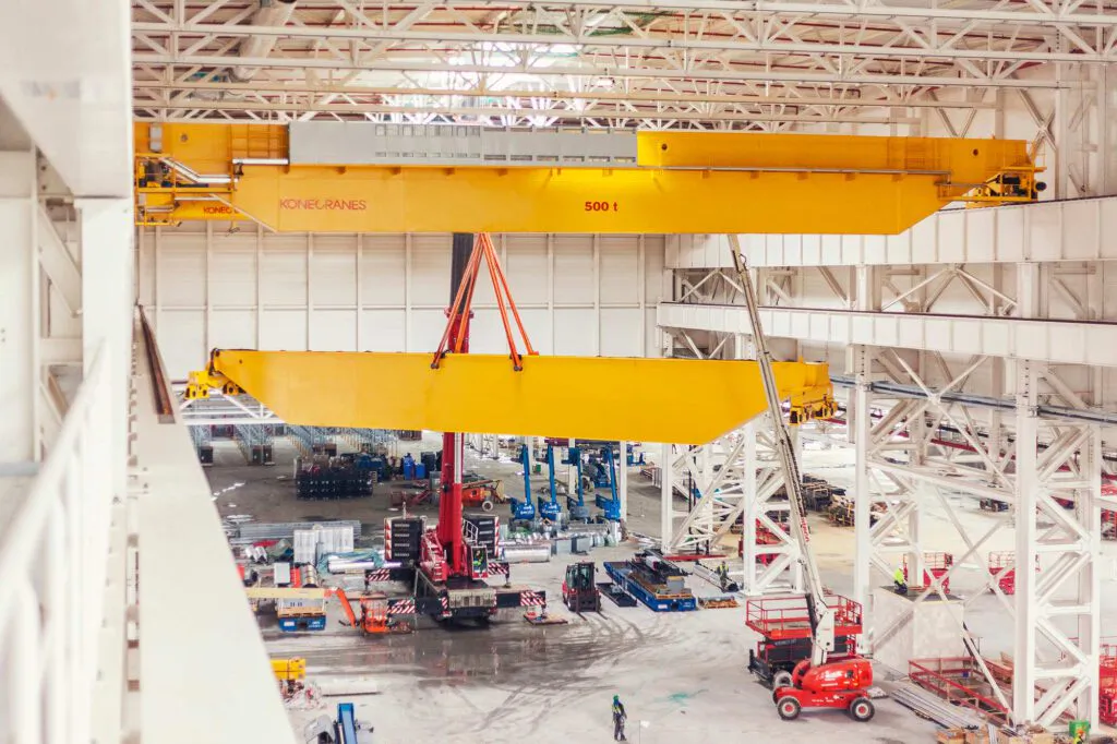 Gelber Konecranes-Brückenkran hebt einen gelben Stahlträger in einer hellen Werkhalle mit Hubarbeitsbühnen.